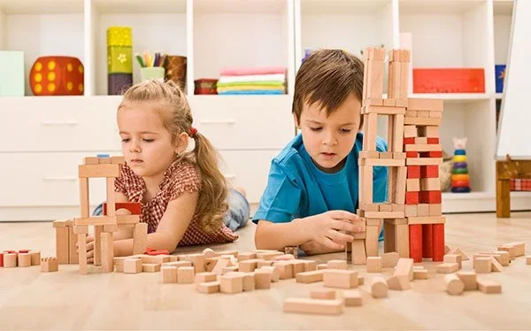 Two children playing with wooden blocks on the floor in a playroom.
