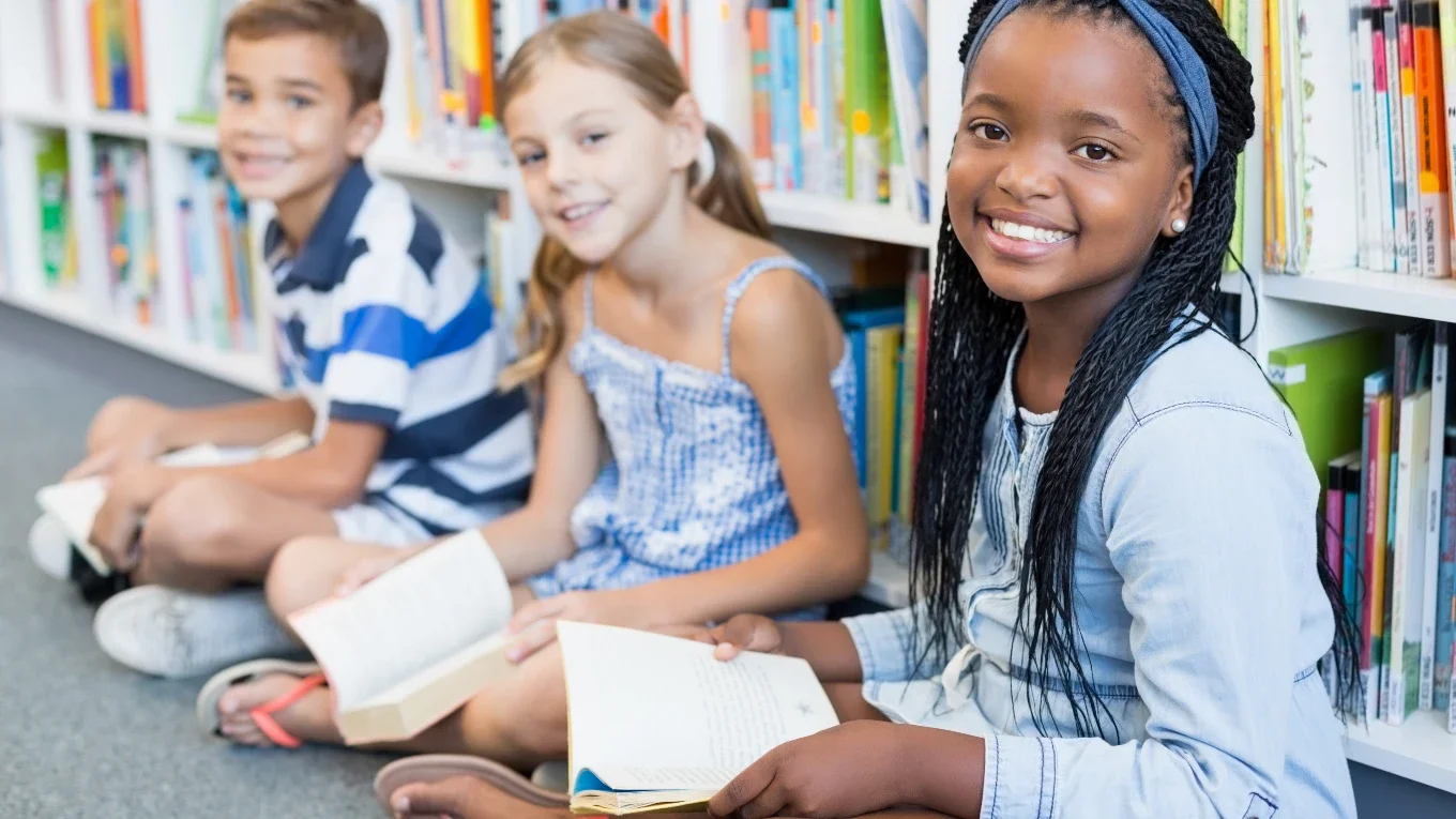 Three children sitting on the floor in a library, holding books and smiling.