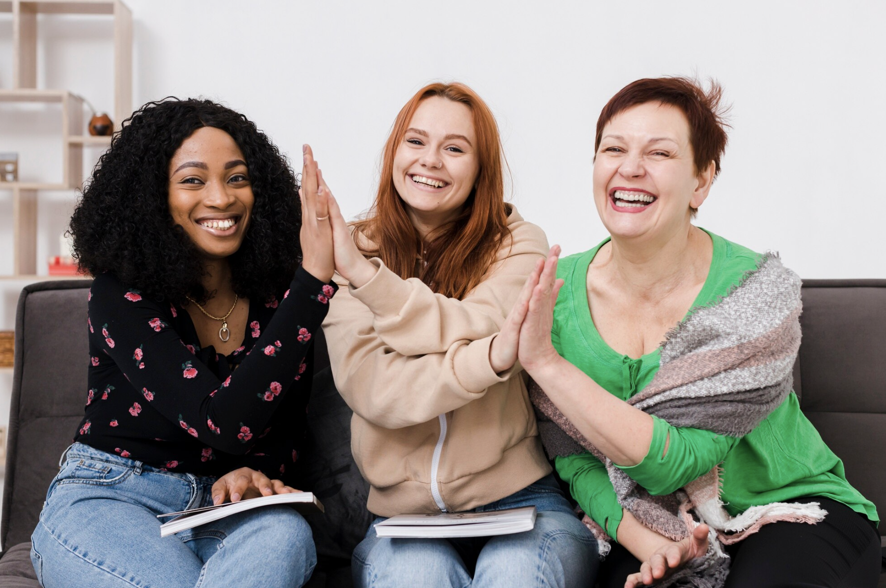 Three women sitting on a couch high-fiving each other and smiling, with notebooks in their laps.