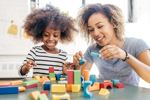 A woman and a young girl playing with colorful wooden building blocks at a table in a bright room.