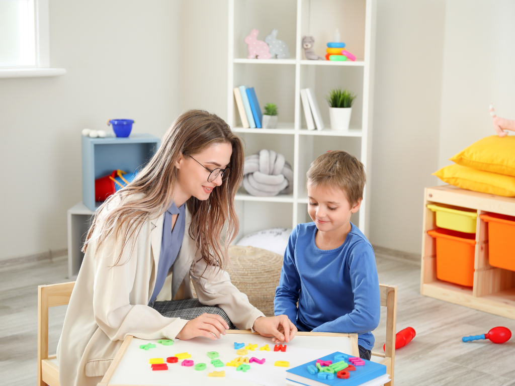A woman and a boy playing with colorful alphabet foam letters at a small table in a children's playroom.