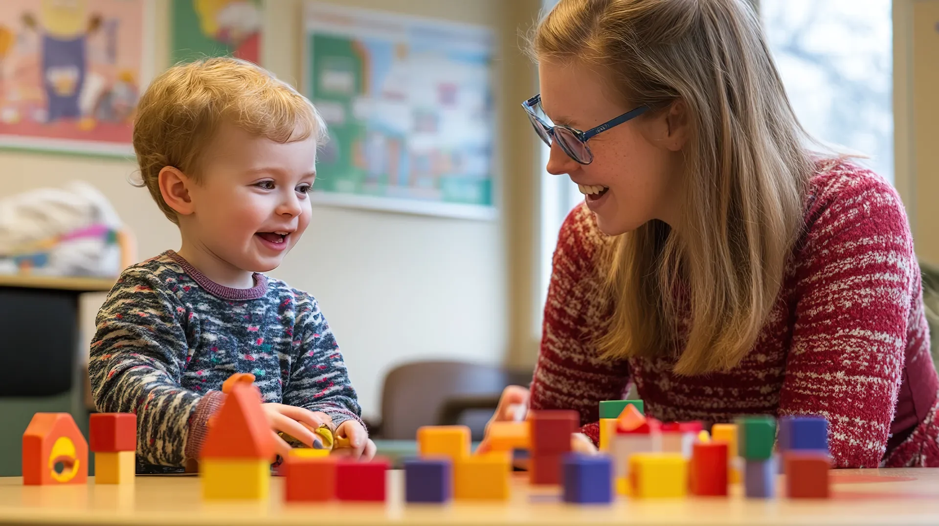 A young boy and a woman, possibly a teacher or caregiver, playing with colorful wooden building blocks at a table in a classroom.