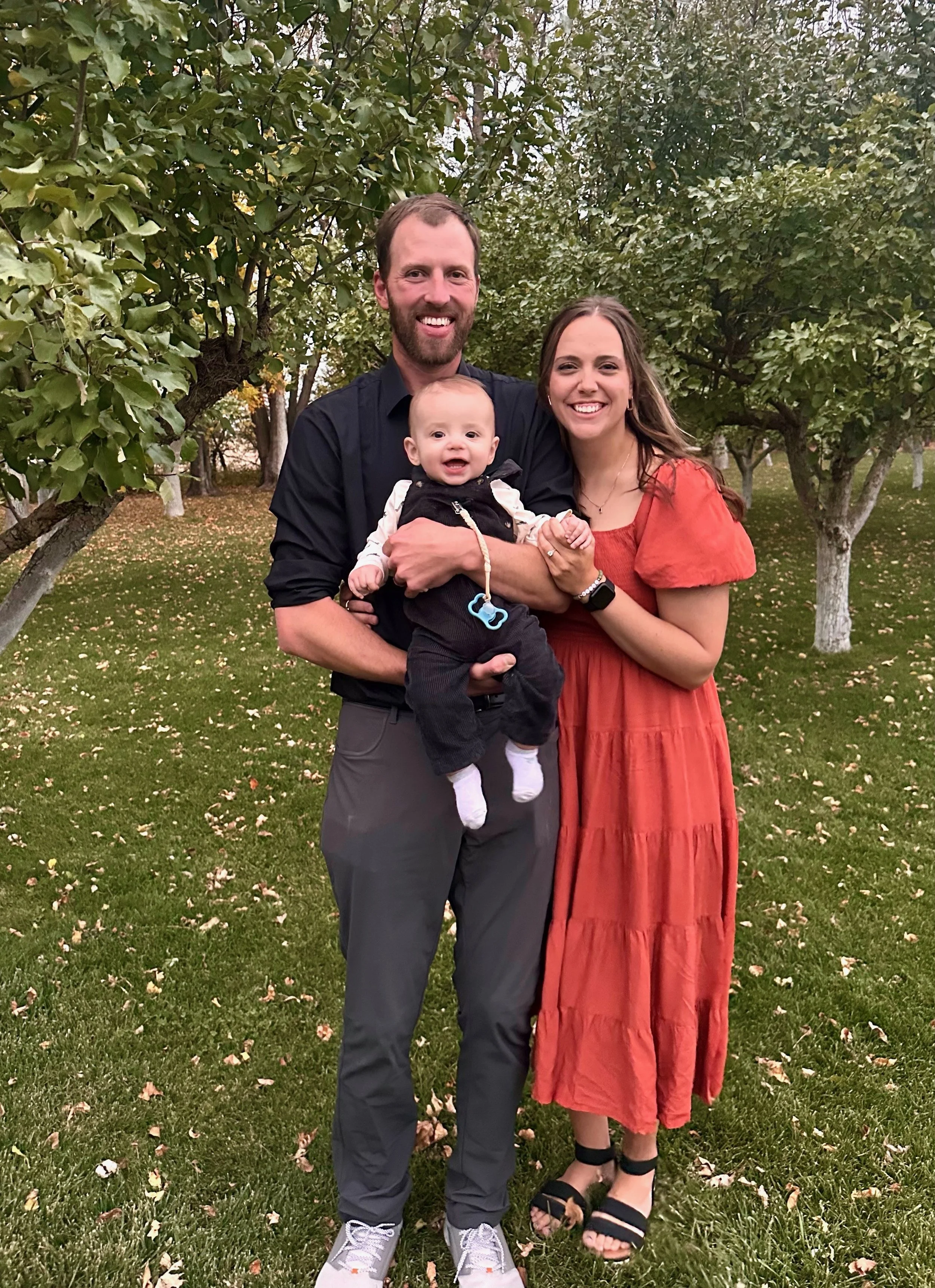 A happy family of three standing on a grassy lawn with trees in the background. The man is holding a baby, and the woman is standing next to them, all smiling at the camera.