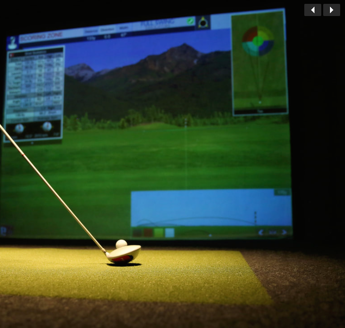 An indoor golf simulator setup showing a golf ball on a tee on a green mat, with a large screen displaying a virtual golf course and mountain scenery in the background.