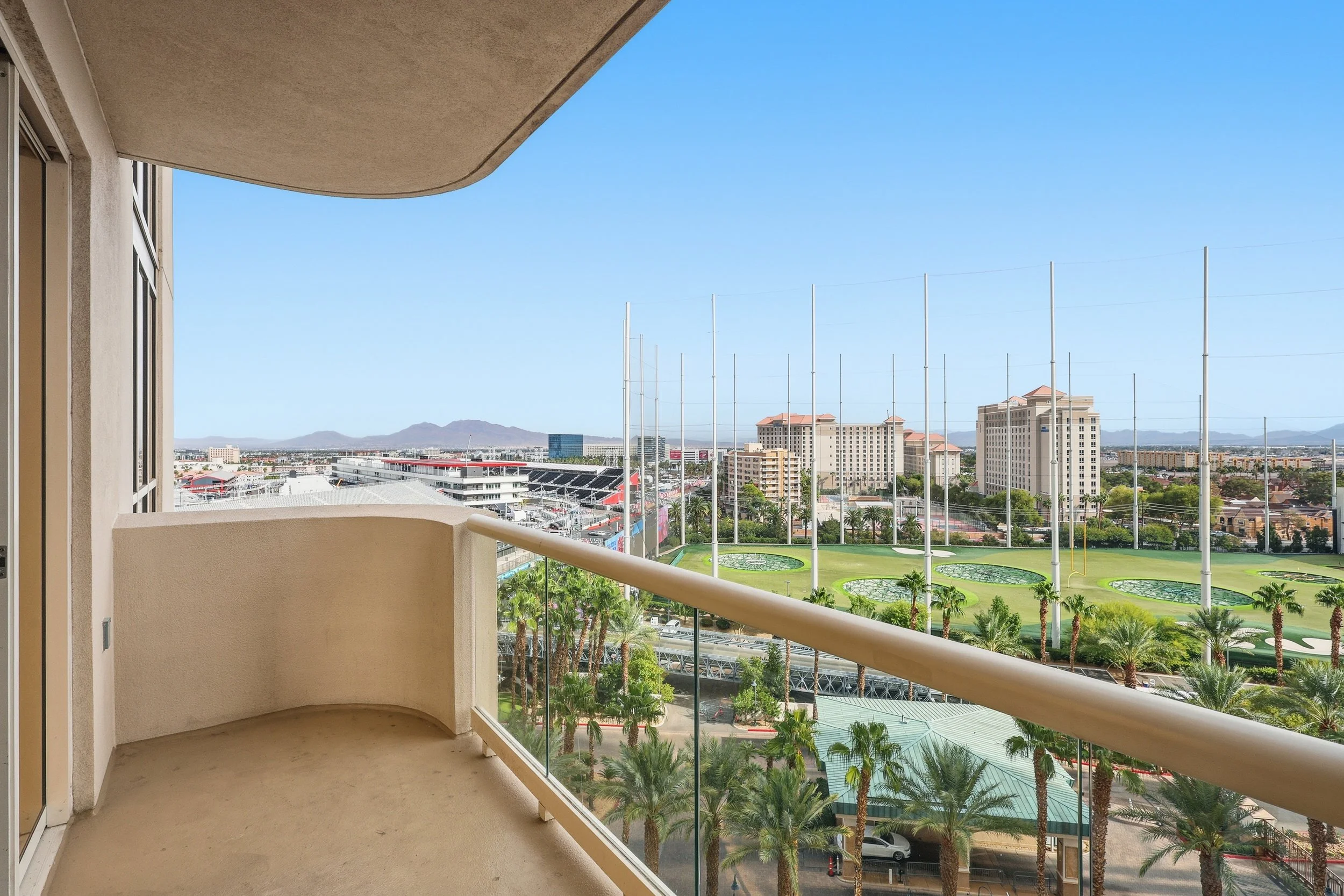 Balcony view overlooking a cityscape with high-rise buildings, palm trees, and a sports stadium with tall poles, under a clear blue sky.