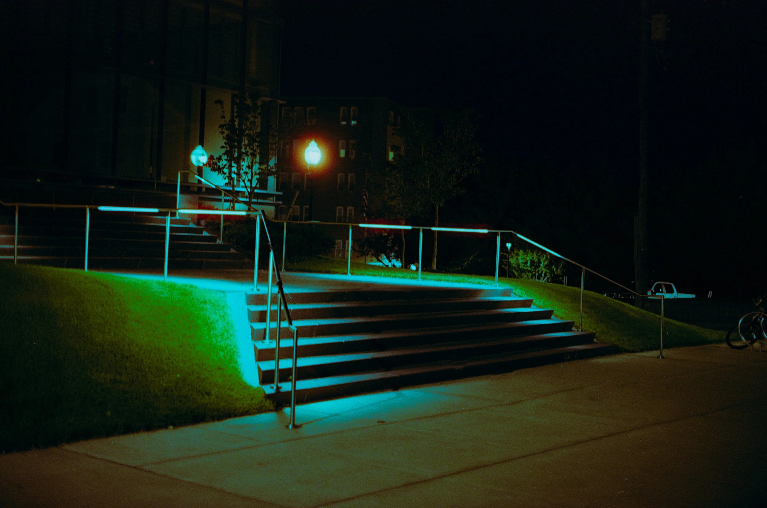 Nighttime scene of an outdoor staircase with handrails, illuminated by colored lights, leading up to a building. The area is dark with some streetlights and a few cars visible in the background.