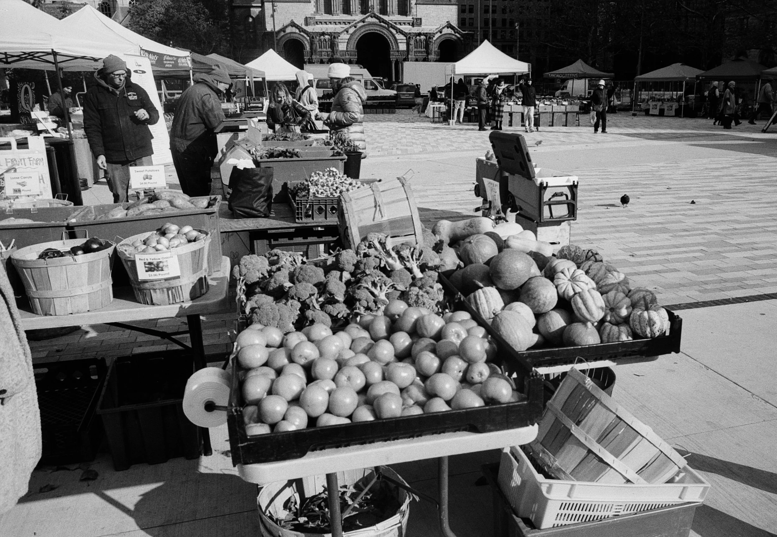 Outdoor farmers market with vendors selling produce like onions, tomatoes, pumpkins, and broccoli. Customers browse and shop, set against historic buildings and tents.