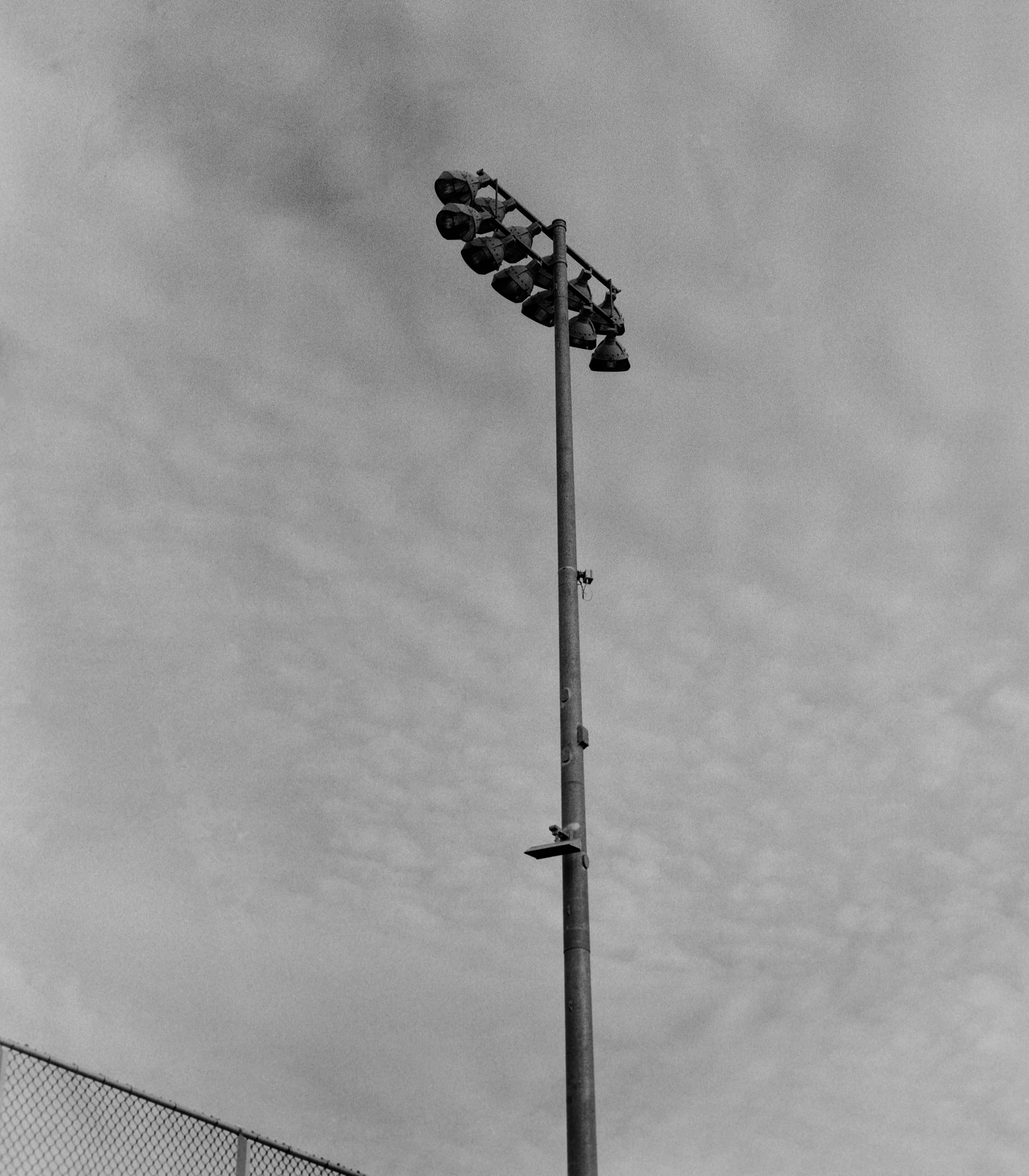 A tall stadium light pole with multiple lights attached, against a cloudy sky, at a sports field, with a chain-link fence in the lower left corner.