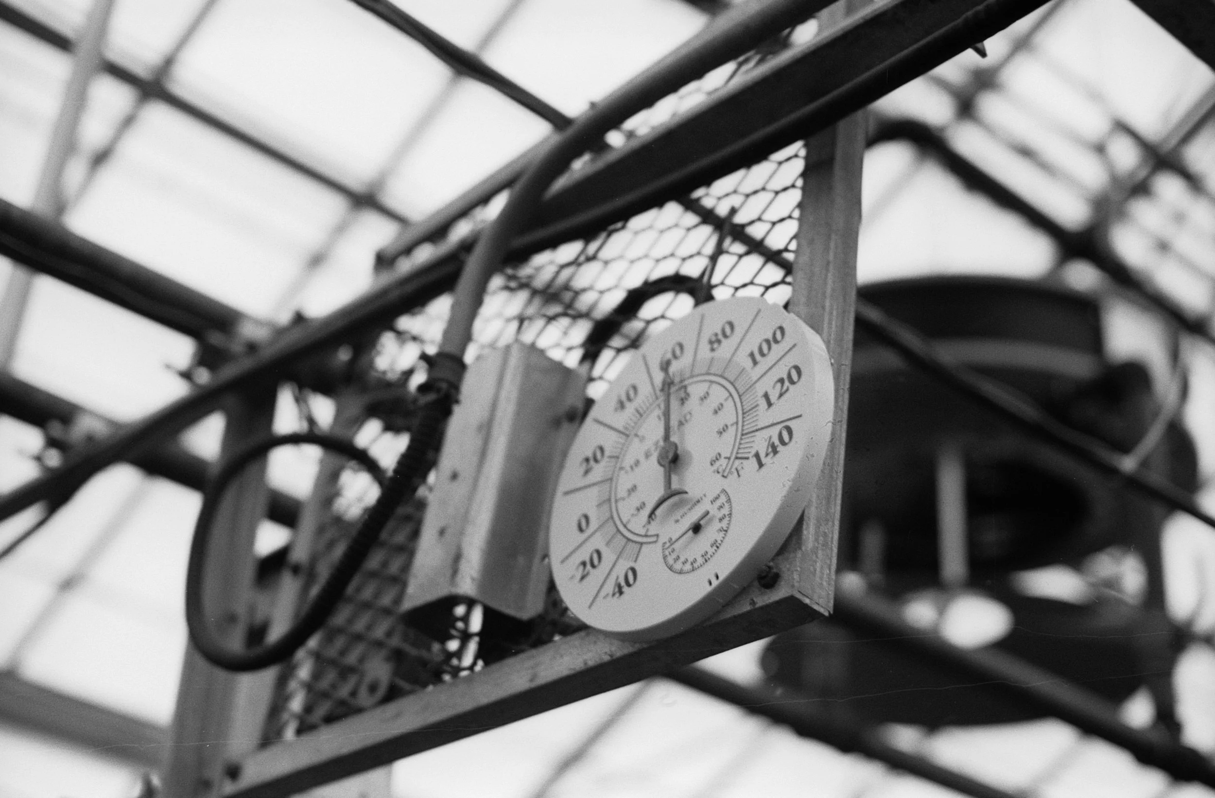 A black and white photograph of a thermometer attached to a wooden structure, with a metal framework and a grid-patterned background, measuring temperature in degrees Fahrenheit.