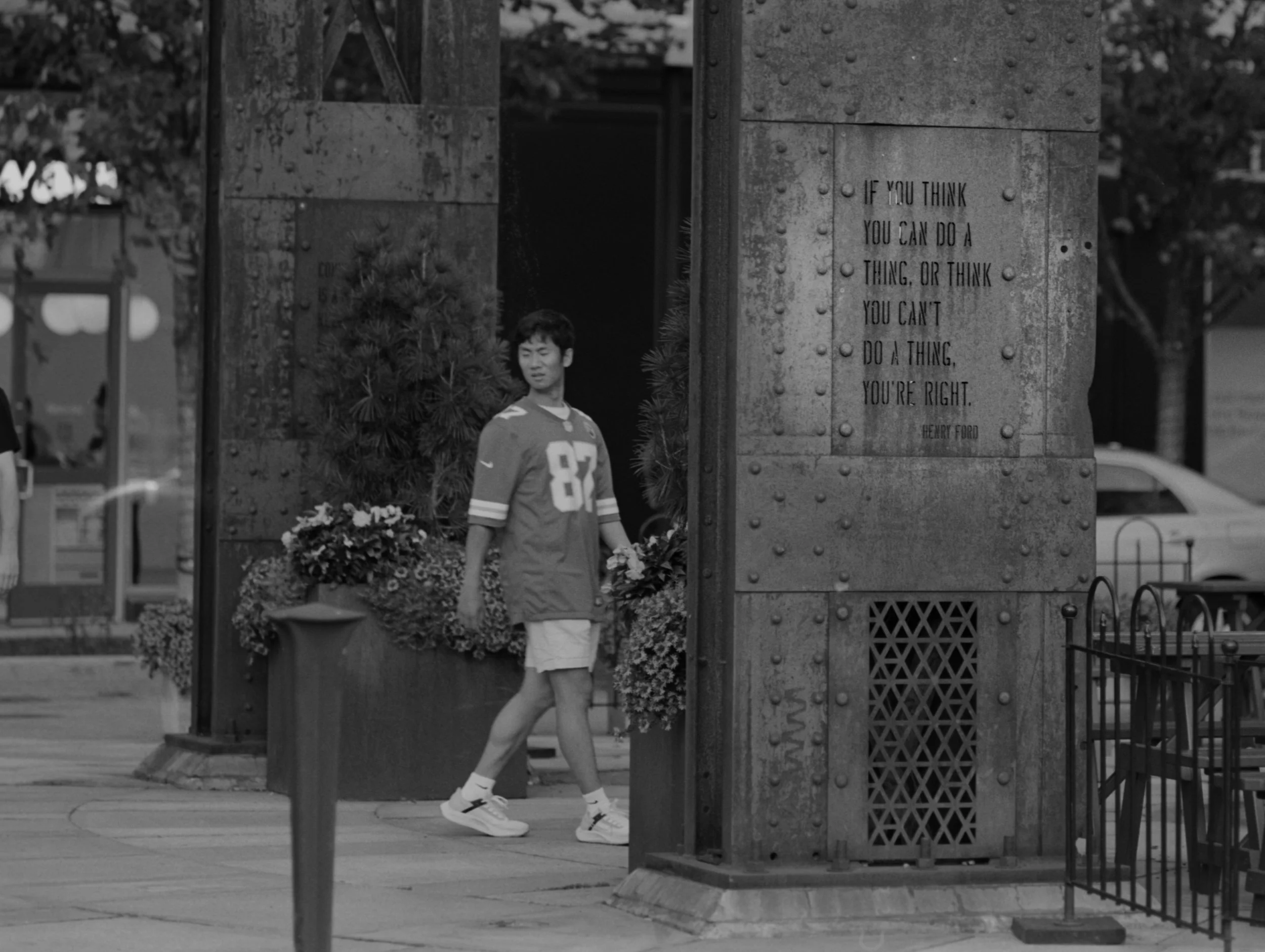 A young man in a sports jersey and shorts walking past a large metal structure with an inspirational quote, while holding flowers in his hand, on a city street.