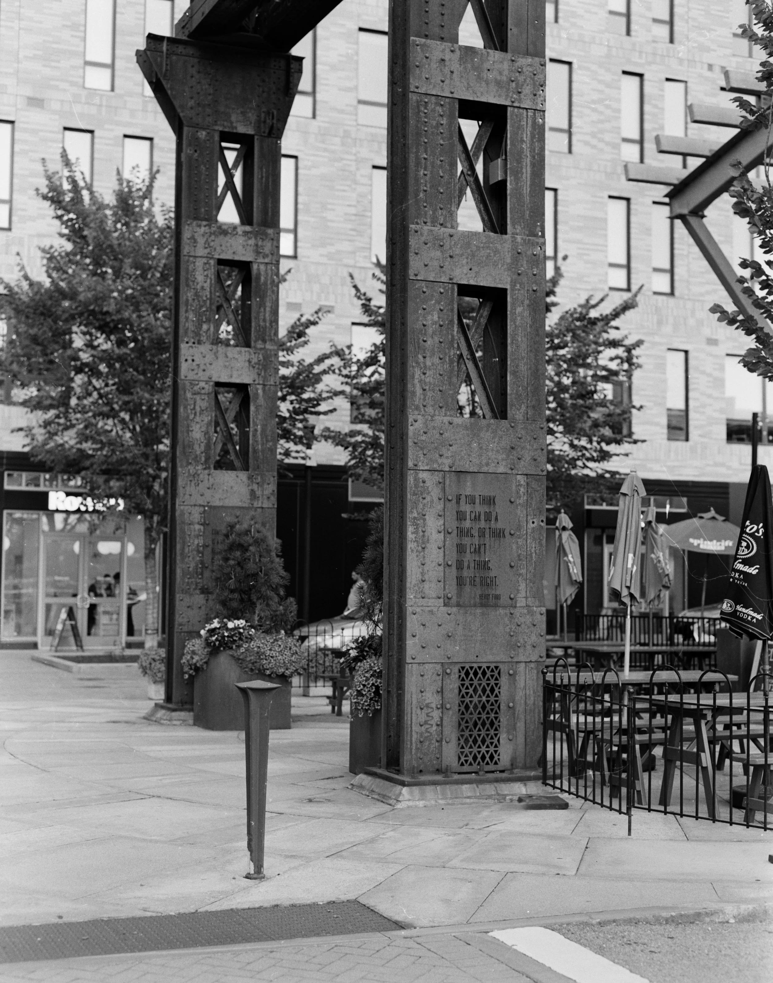 Urban outdoor scene featuring a large metal sculpture or structure, trees, a building with multiple windows, and an outdoor seating area with umbrellas and chairs.