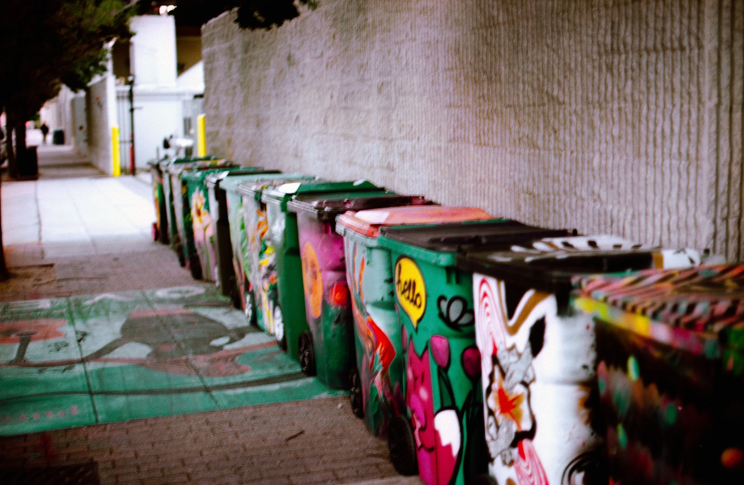 A row of colorful, graffiti-painted trash bins lined up against a beige brick wall along a sidewalk.