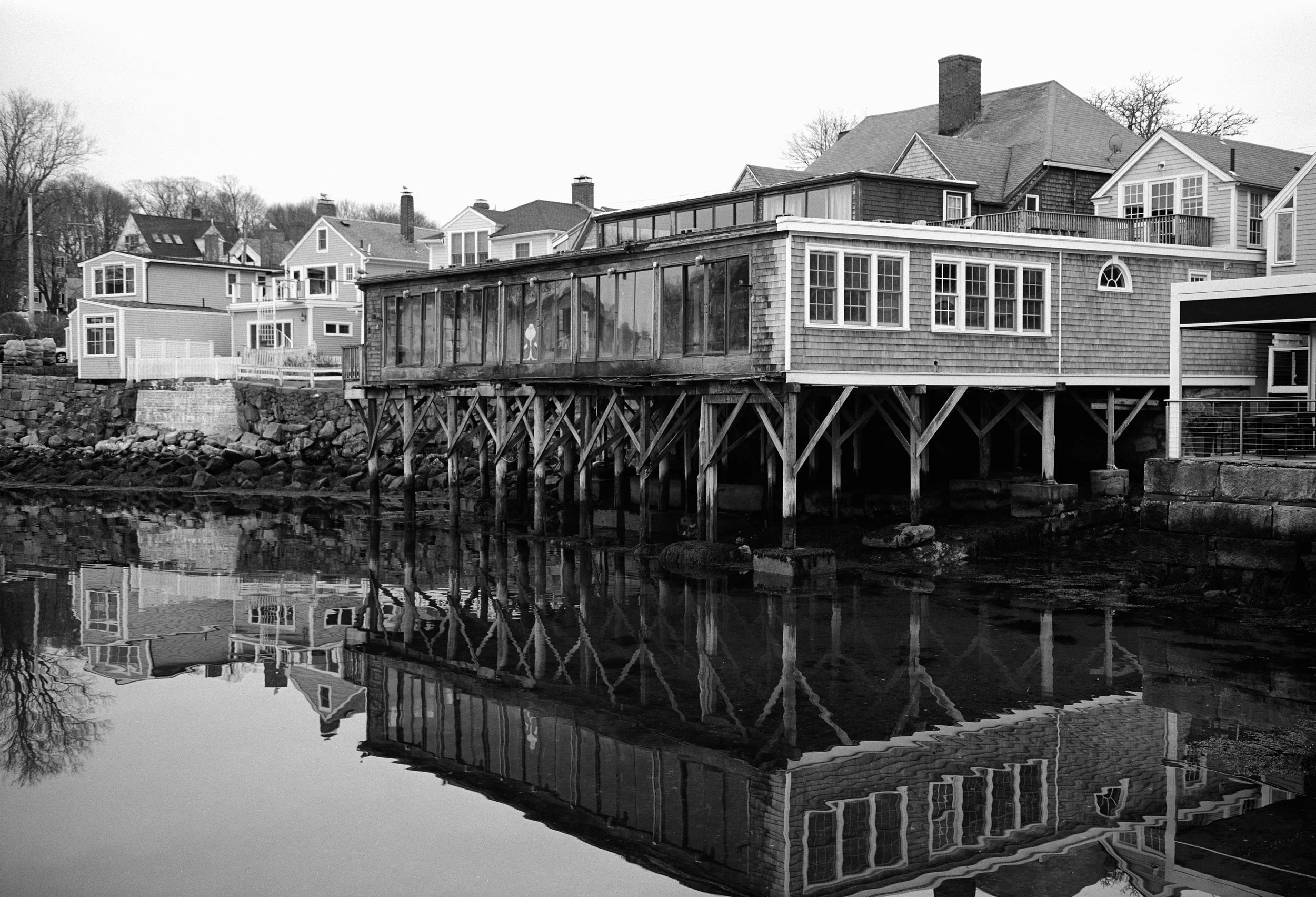 A black and white photo of a waterfront house with a wooden deck on stilts, with multiple windows, and neighboring houses along the shoreline.
