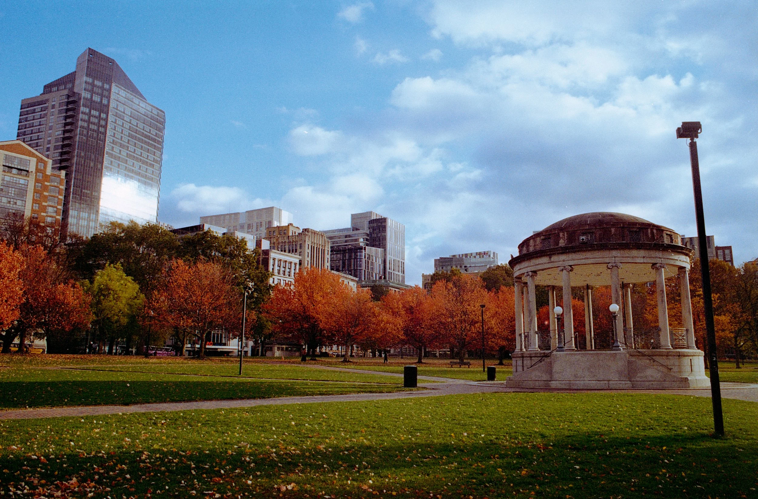 City park with trees displaying fall foliage, a pavilion with columns, and tall buildings under a partly cloudy sky.
