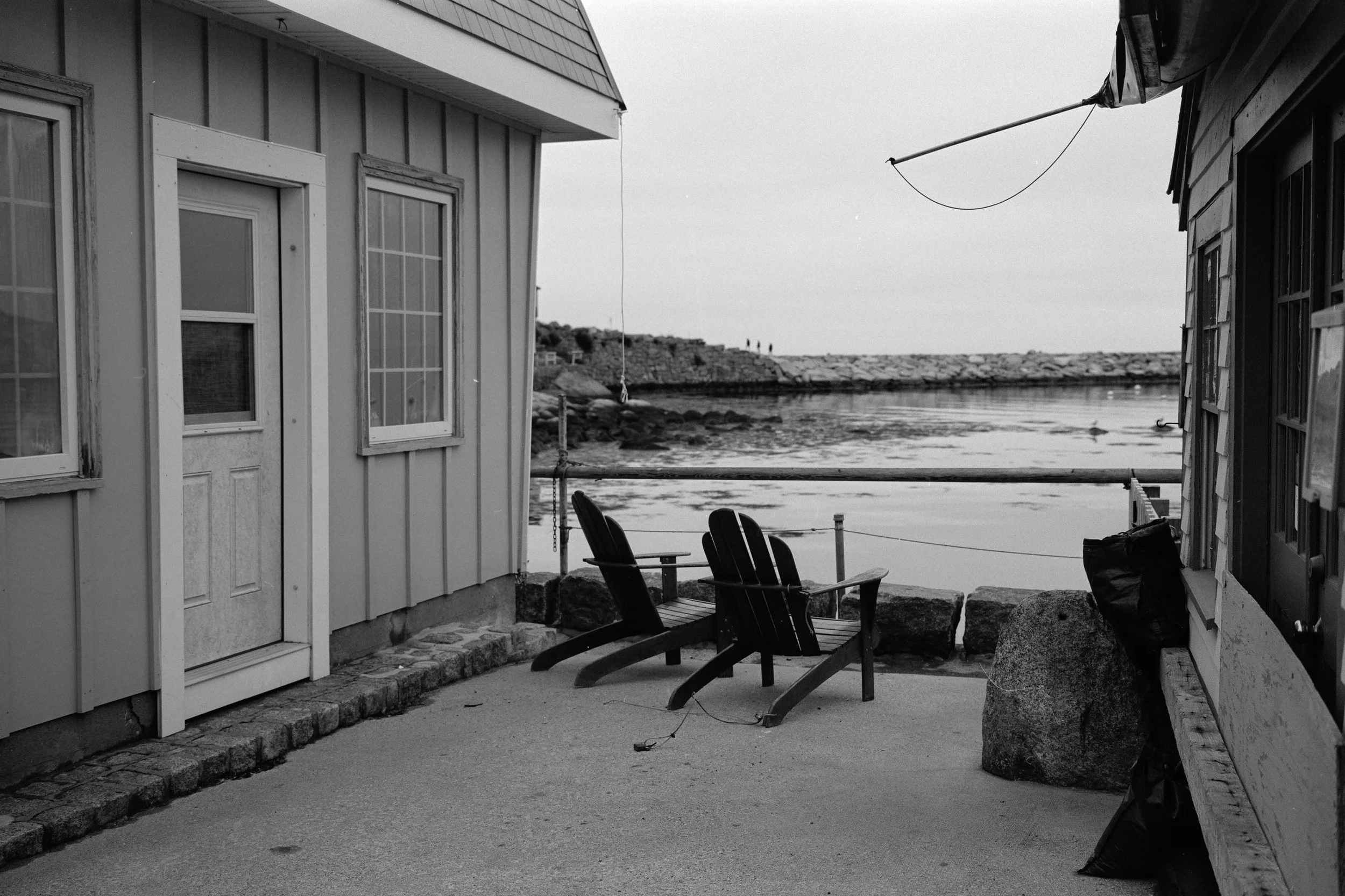 Two wooden Adirondack chairs on a patio facing a body of water, with a rocky shoreline and two people walking in the distance.