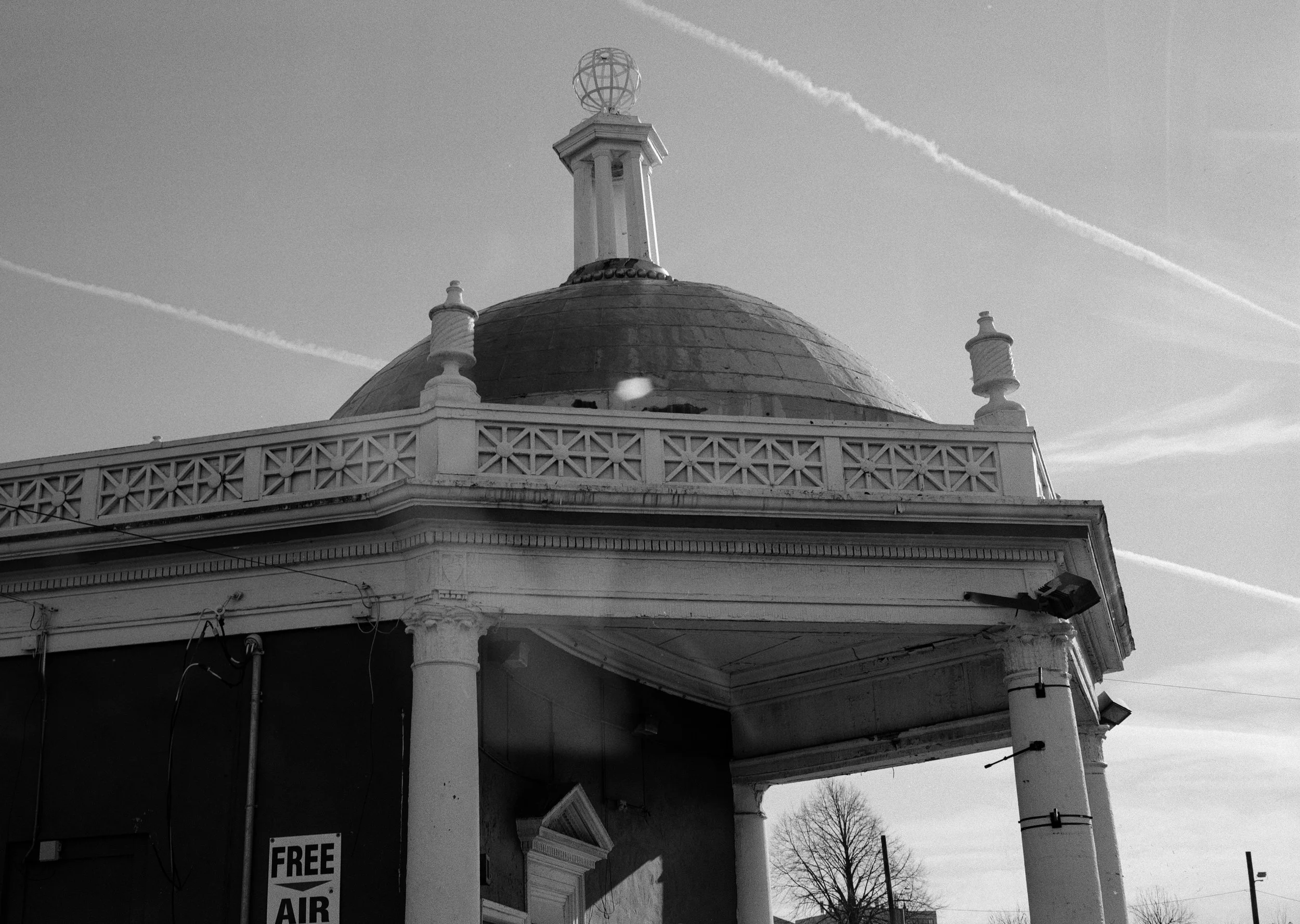 Black and white photo of an old building with a domed roof, columns, and decorative railing, with airplanes contrails in the sky.