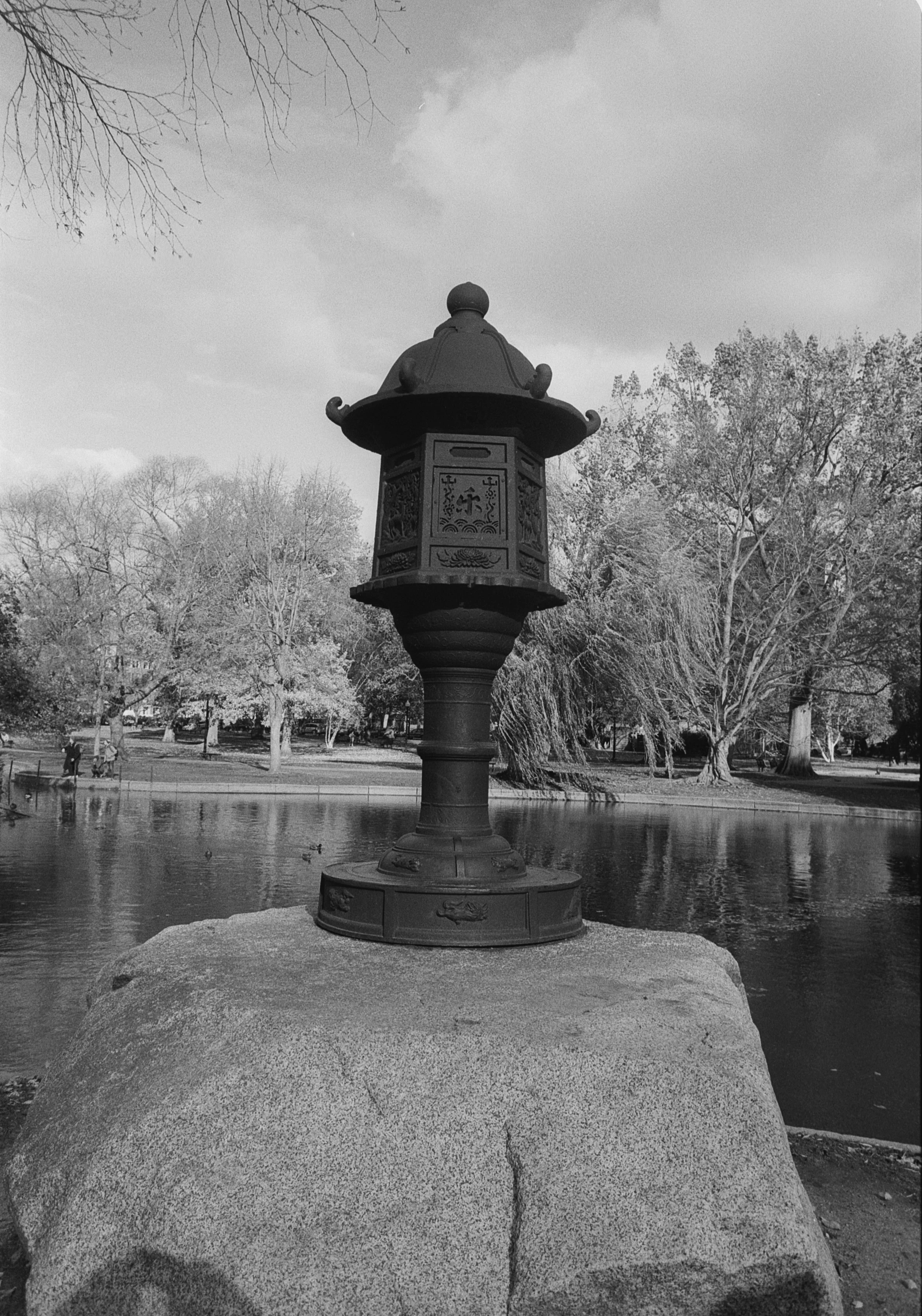 A black and white photo of a traditional Asian-style lantern placed on a large rock near a pond in a park. The background shows trees with some bare branches and water with ducks swimming.