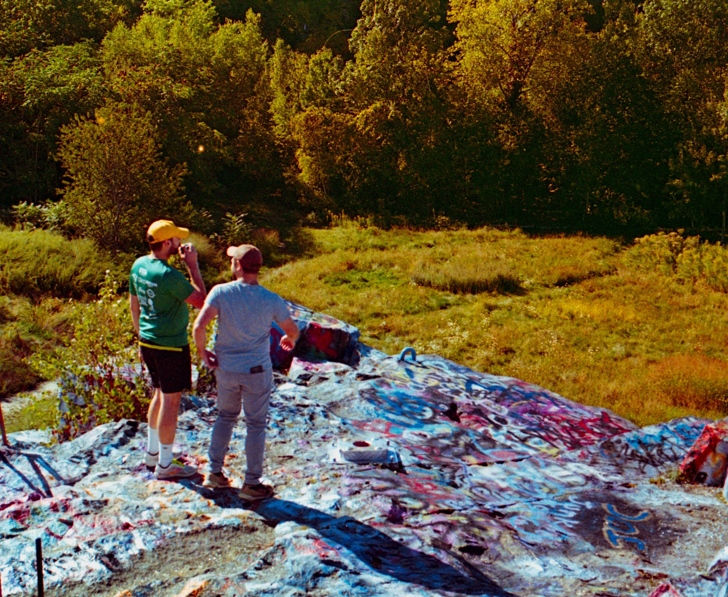 Two men standing on a large graffiti-covered tarp in a grassy field with trees in the background, one drinking from a bottle.
