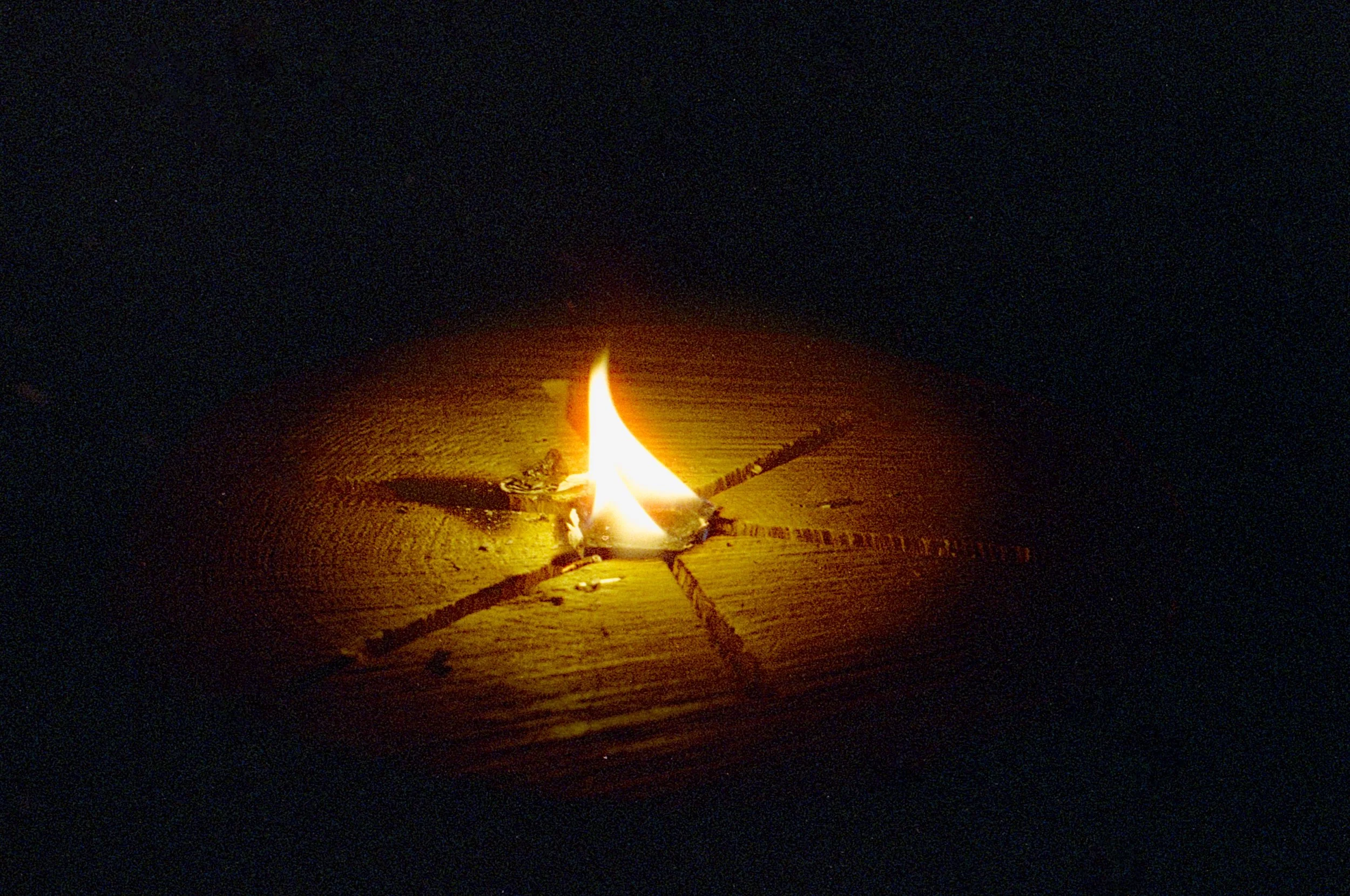 A lit candle with a bright flame resting on a wooden surface in a dark environment.