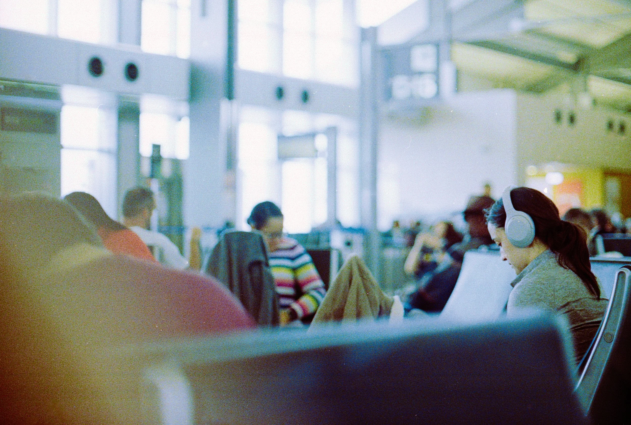 People sitting in an airport waiting area, with a woman in a gray sweater wearing headphones in the foreground, others are visible in the background.