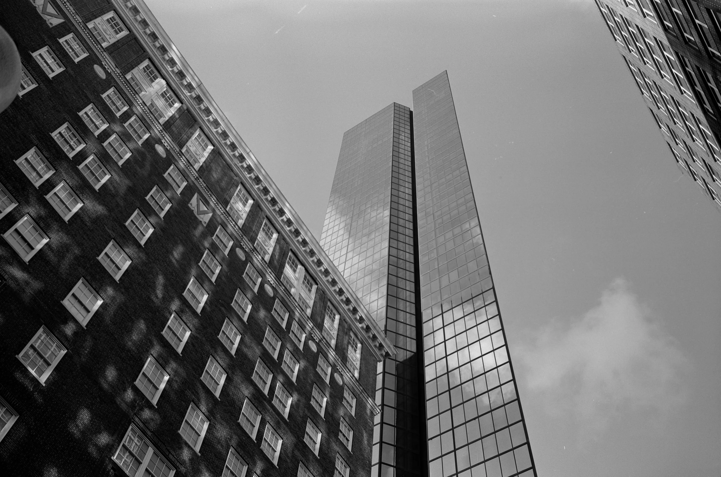 Black and white photo of city skyscrapers taken from ground level looking up, with a modern glass tower and older brick building.