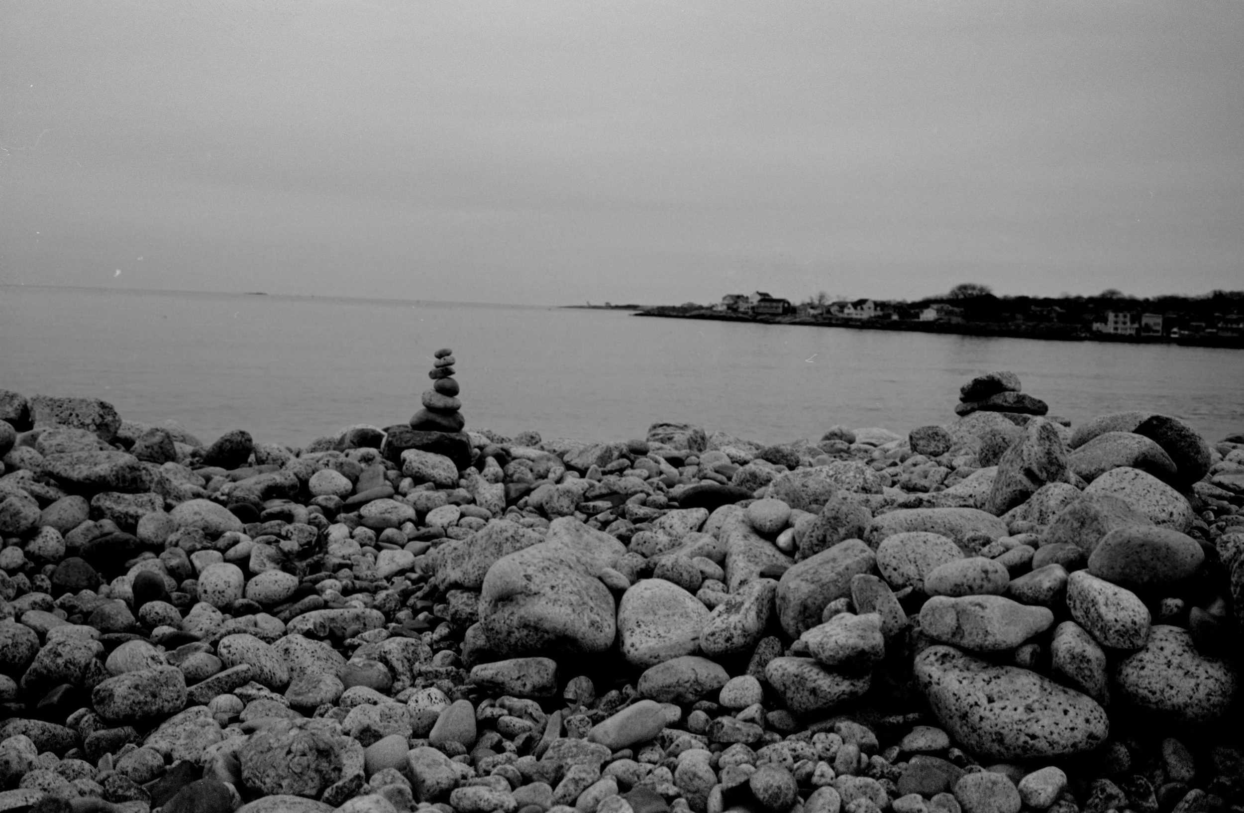 A black and white photo of a rocky beach with two stacked stone cairns in the foreground, a calm body of water, and houses on the distant shoreline.