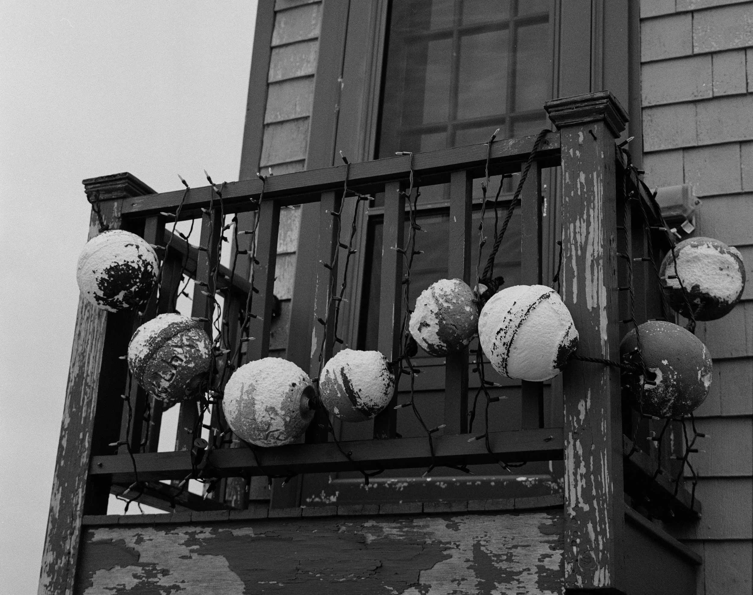 A weathered balcony with peeling paint, decorated with snow-covered Christmas lights and spherical ornaments, next to a window on a house with wooden siding.