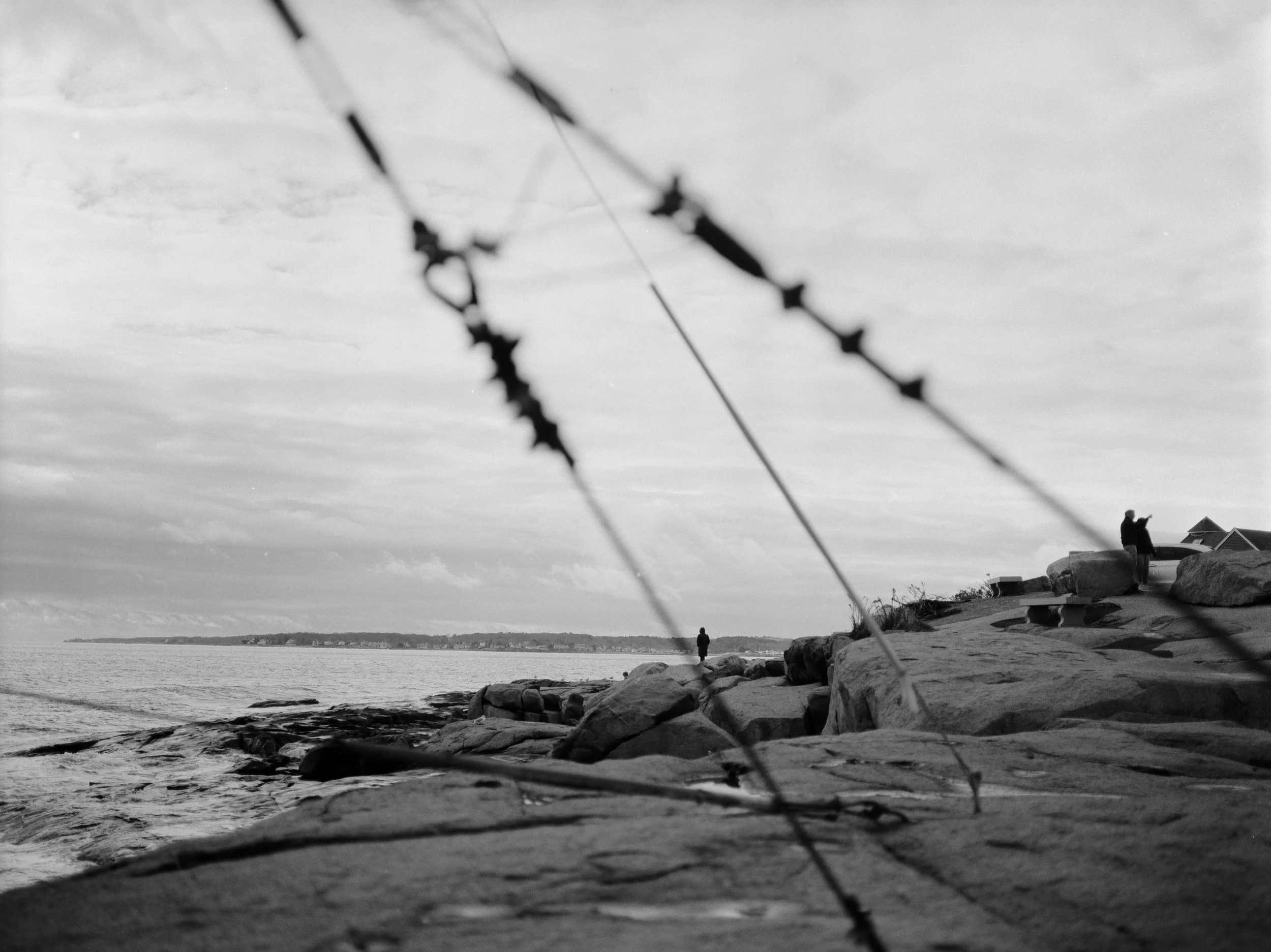 Black and white photo of a rocky shoreline with a person standing near the water and two people sitting on rocks, with a cloudy sky overhead.