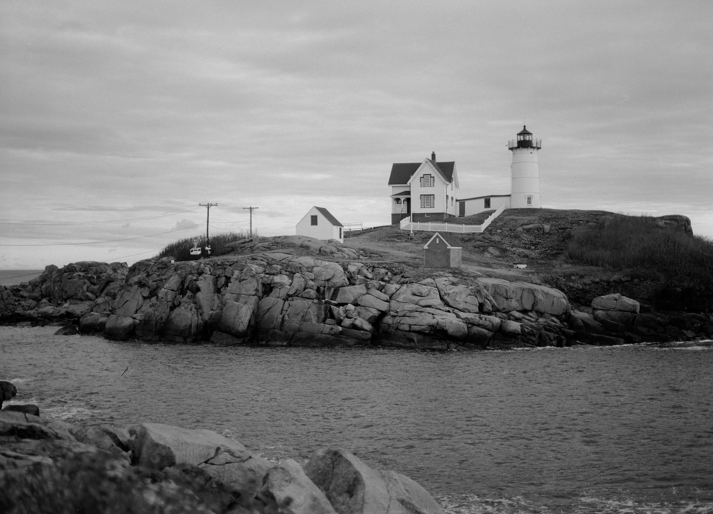 A black and white photo of a lighthouse on a rocky coastline with a house and small building nearby, under a cloudy sky.