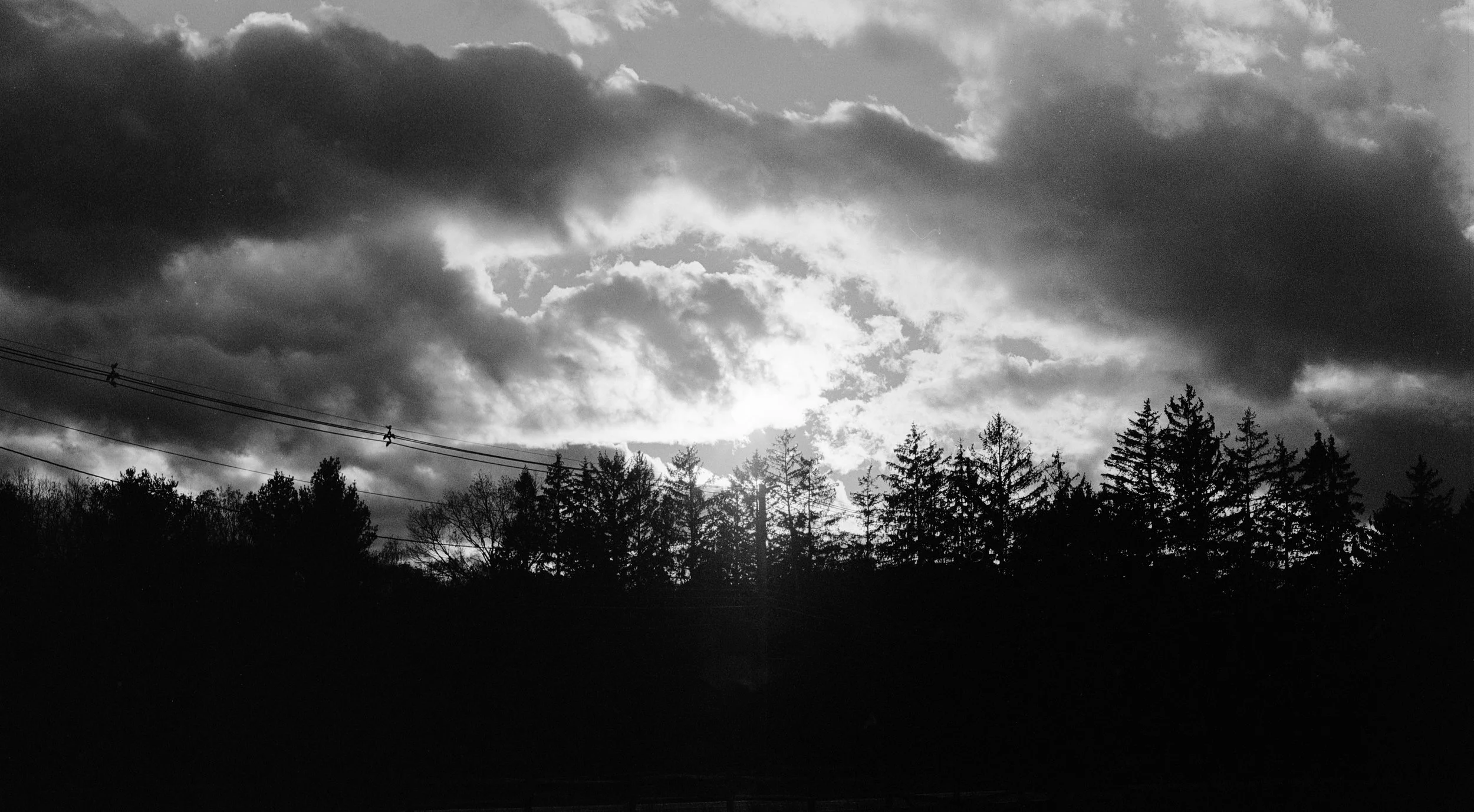 Black and white photo of a cloudy sky with trees and power lines in the foreground.