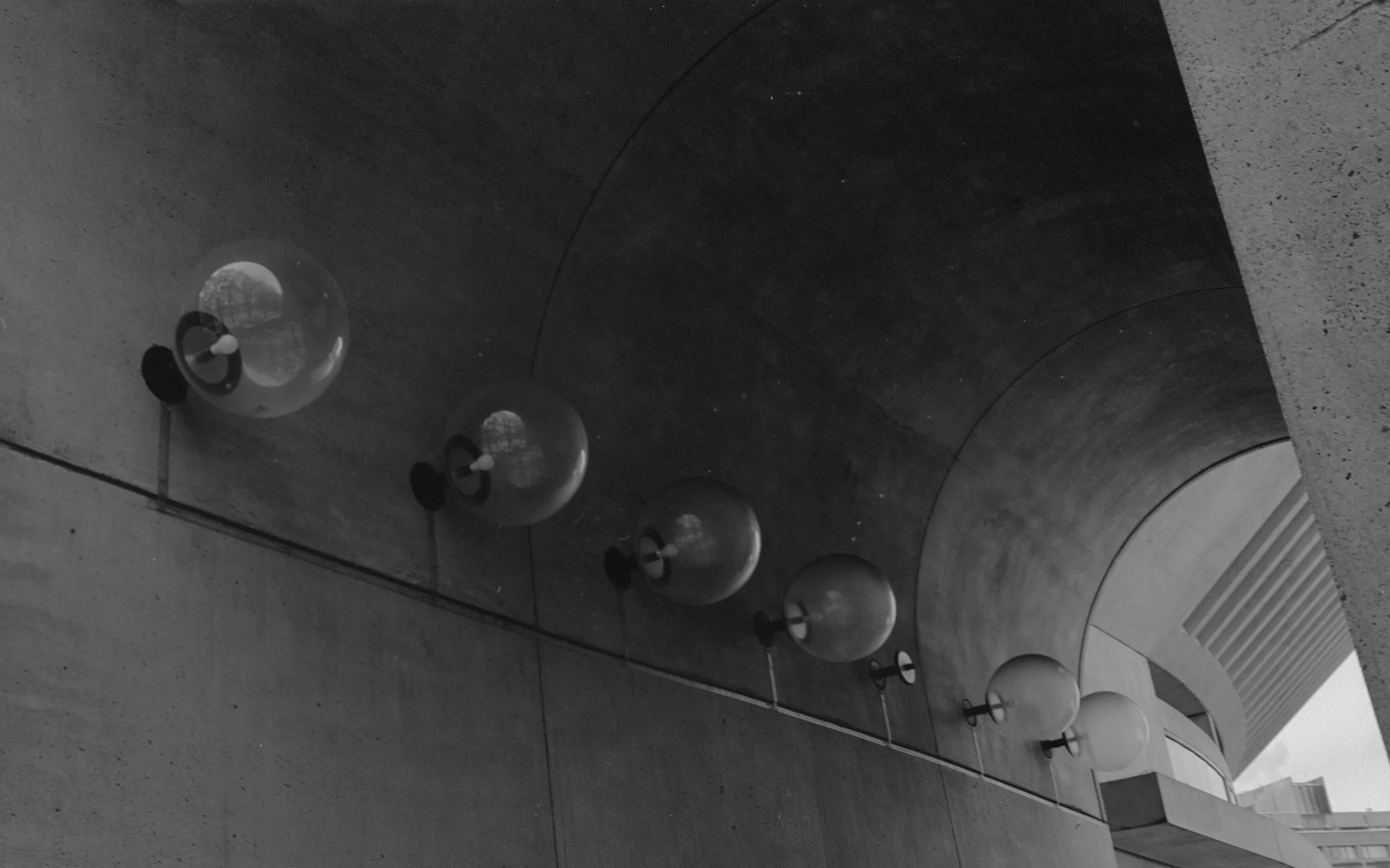 A black-and-white photo of six spherical wall-mounted lamps arranged in a line on a concrete building wall, with a curved architectural feature and a window in the background.