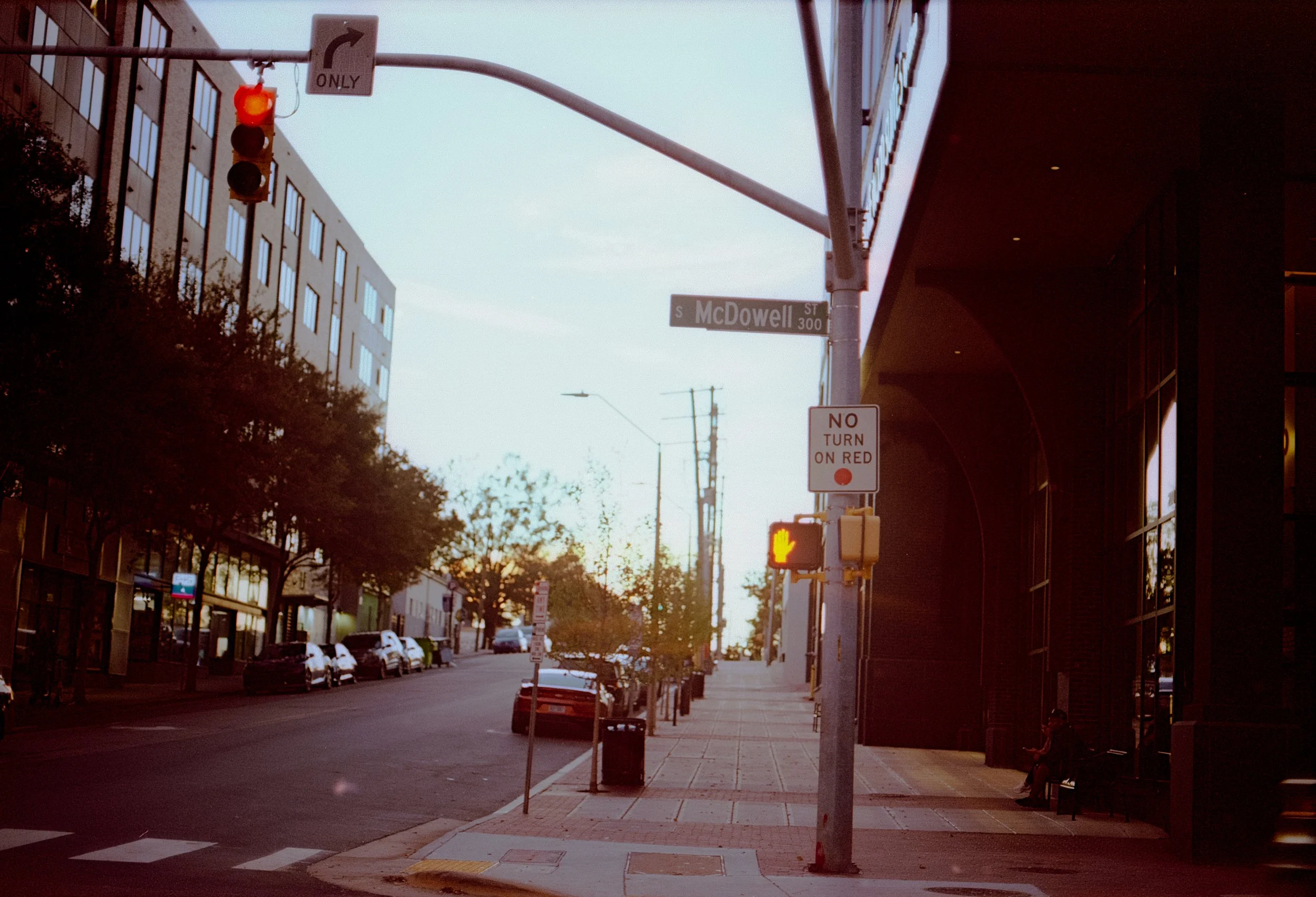 City street corner at sunset with traffic light showing red, street signs reading 'South McDowell' and 'Street 300', parked cars along the street, people sitting on benches, trees lining the sidewalk, and tall buildings on the left.