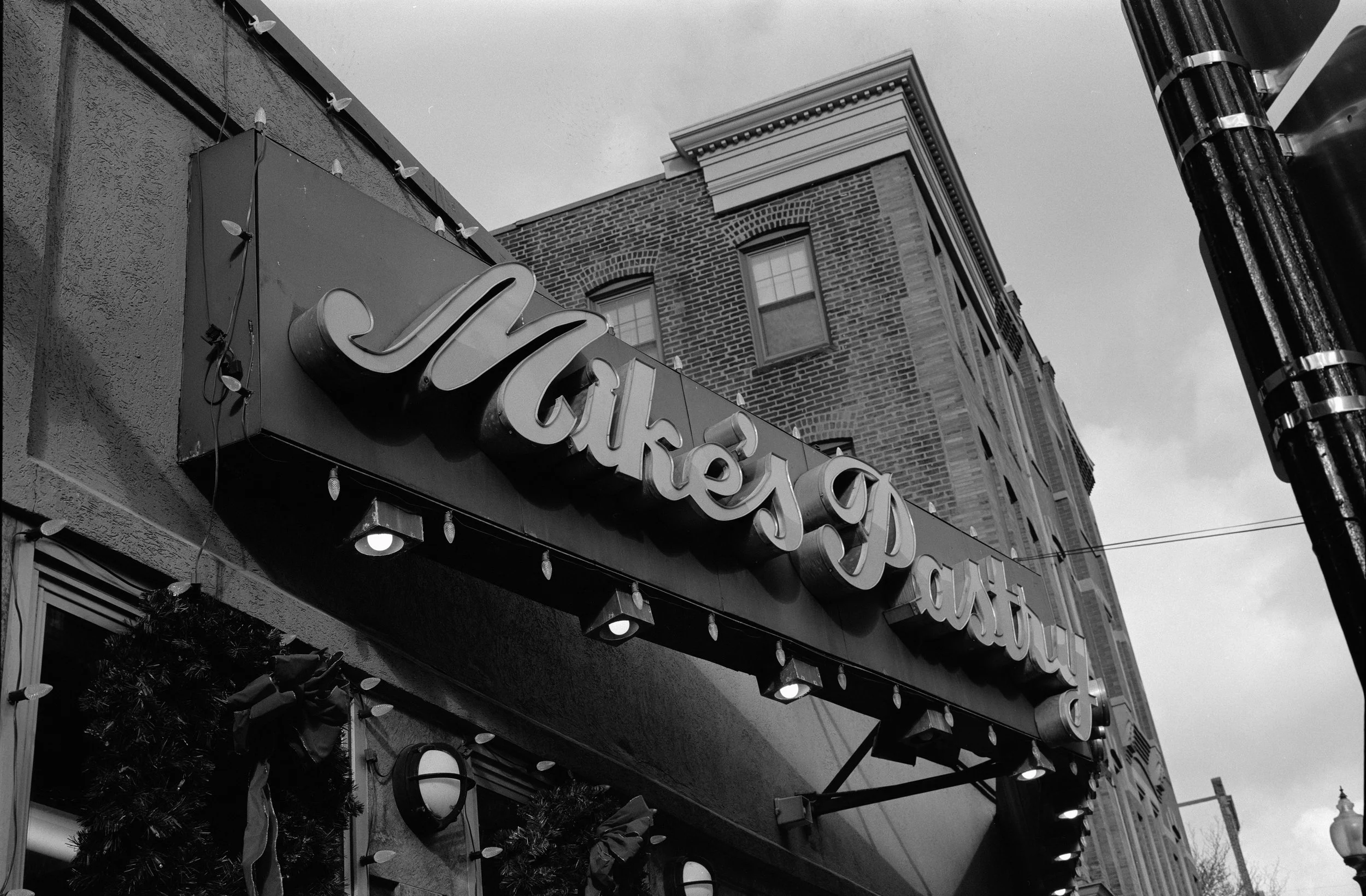 A black-and-white photo of a restaurant with a sign that says 'Makesuparty'. The sign is decorated with string lights, and there is brick and other building architecture visible in the background, along with a cloudy sky.