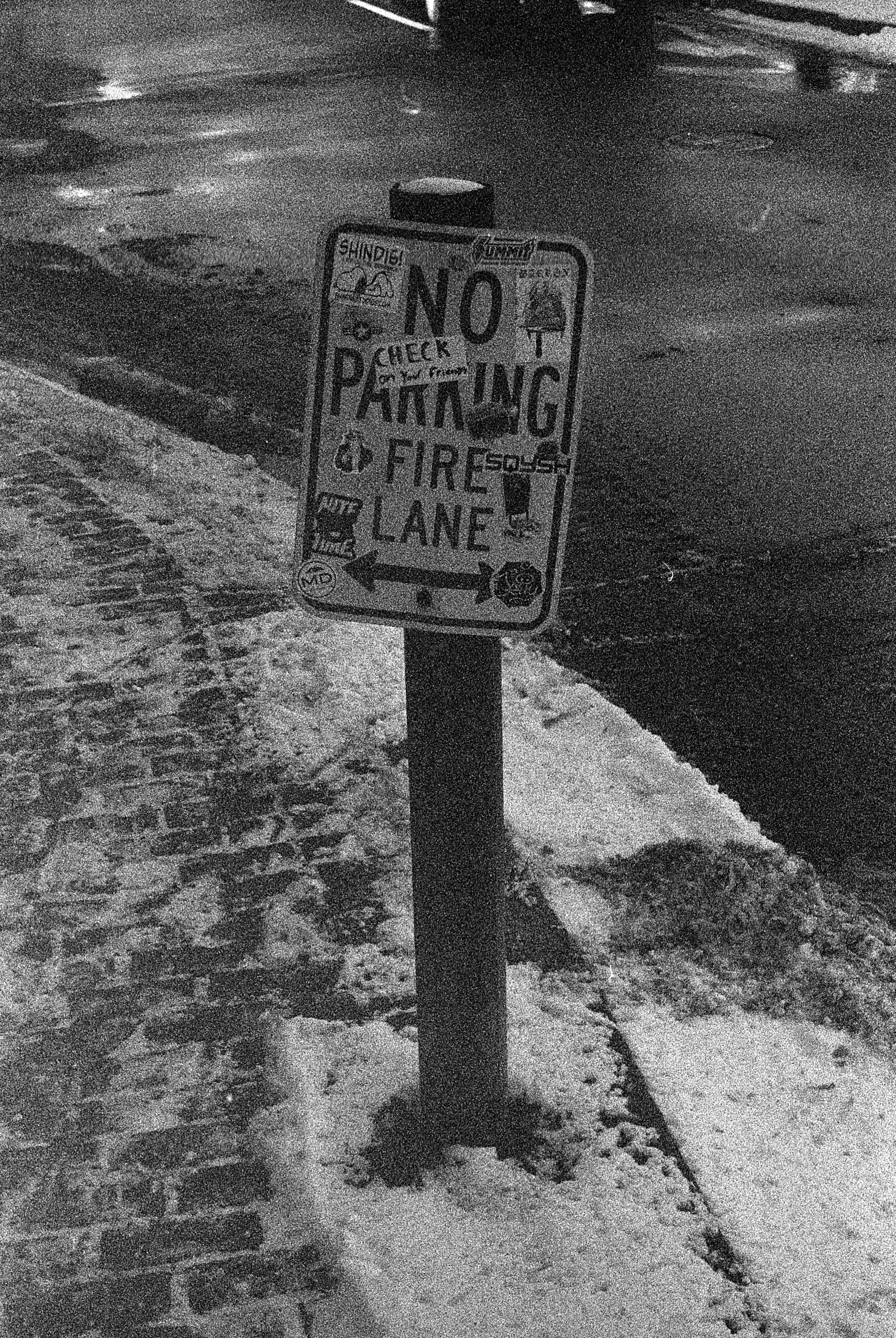 A snow-covered sidewalk and street at night with a sign that says 'No Parking.' The sign has multiple stickers and annotations, and there is visible snow and ice on the ground.