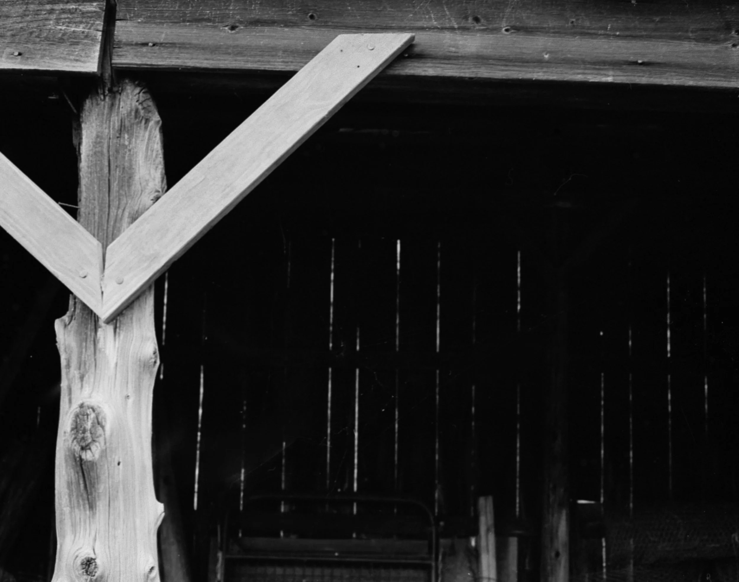 Black and white photograph of a wooden structure, showing a diagonal support beam attached to a vertical post and horizontal beams above. The background is dark with vertical lines, possibly representing wooden panels or a fence.