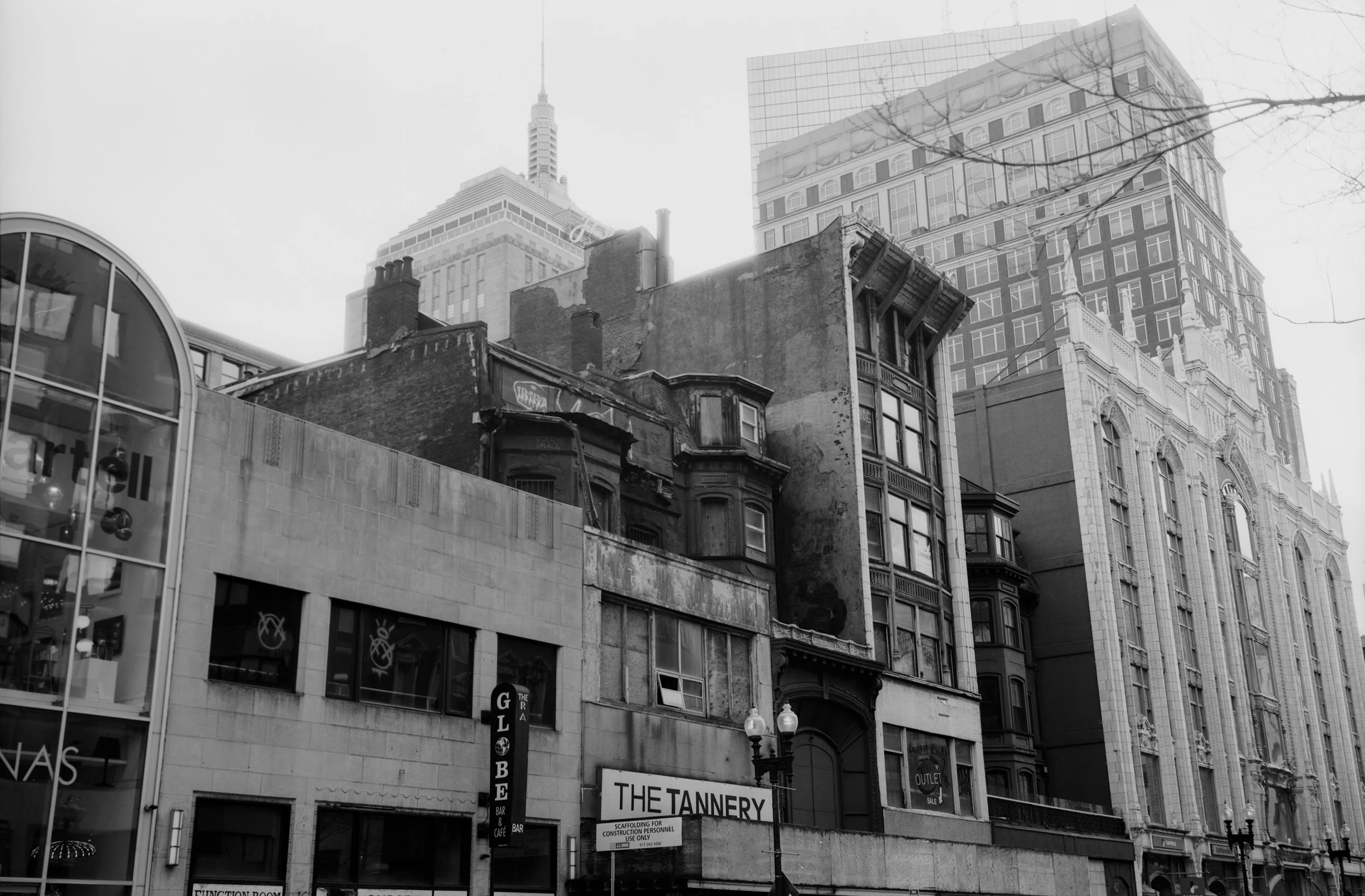 A city street scene with older and modern buildings, including a retail store with a curved glass facade, and signs for a bar and a clothing outlet, with the city skyscrapers in the background.