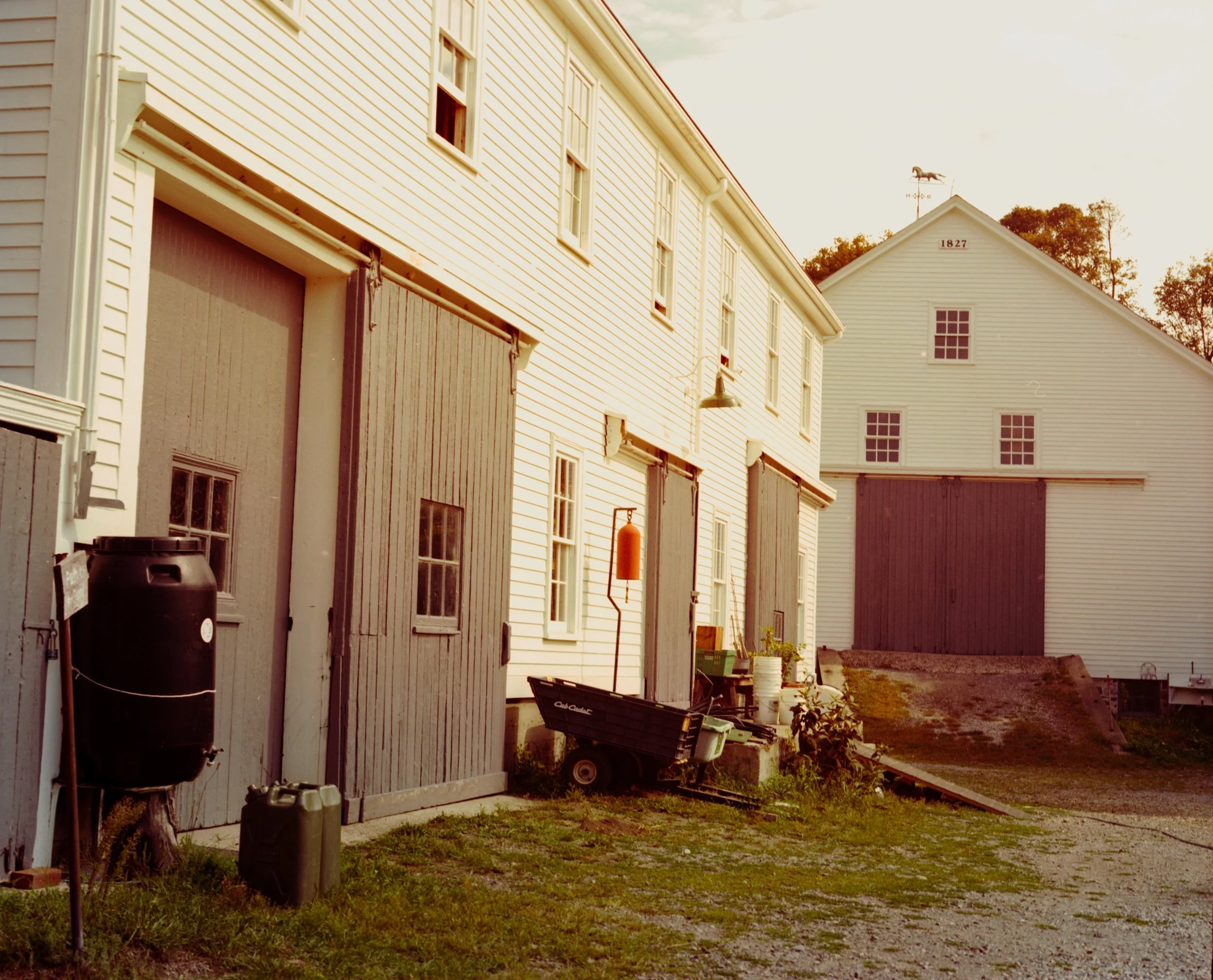 A farm scene with white wooden buildings, a barn with a weather vane, and outdoor farm equipment in the late afternoon or early evening.