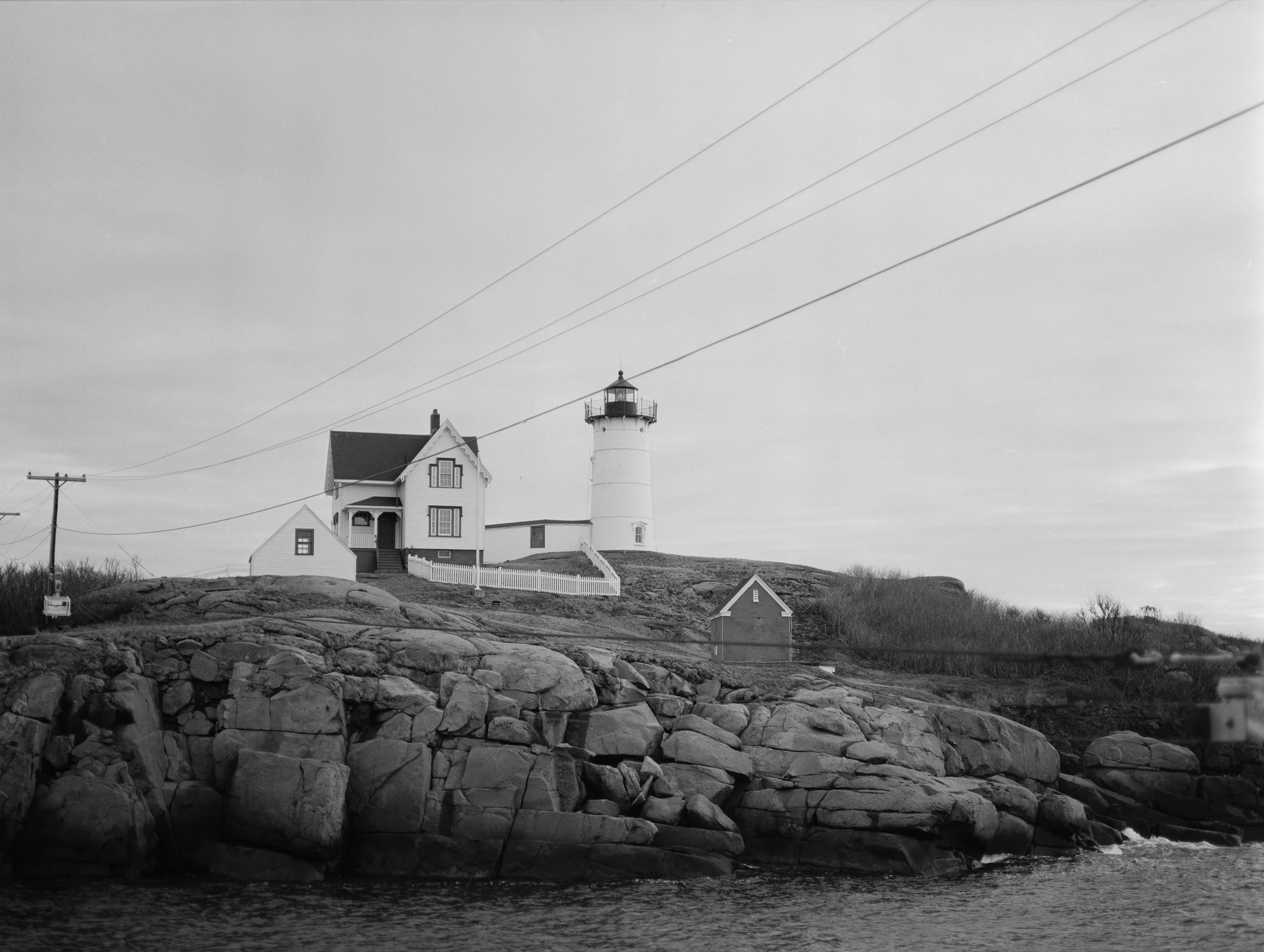 A black and white photograph of a lighthouse and house on a rocky coastline with water in the foreground and power lines overhead.