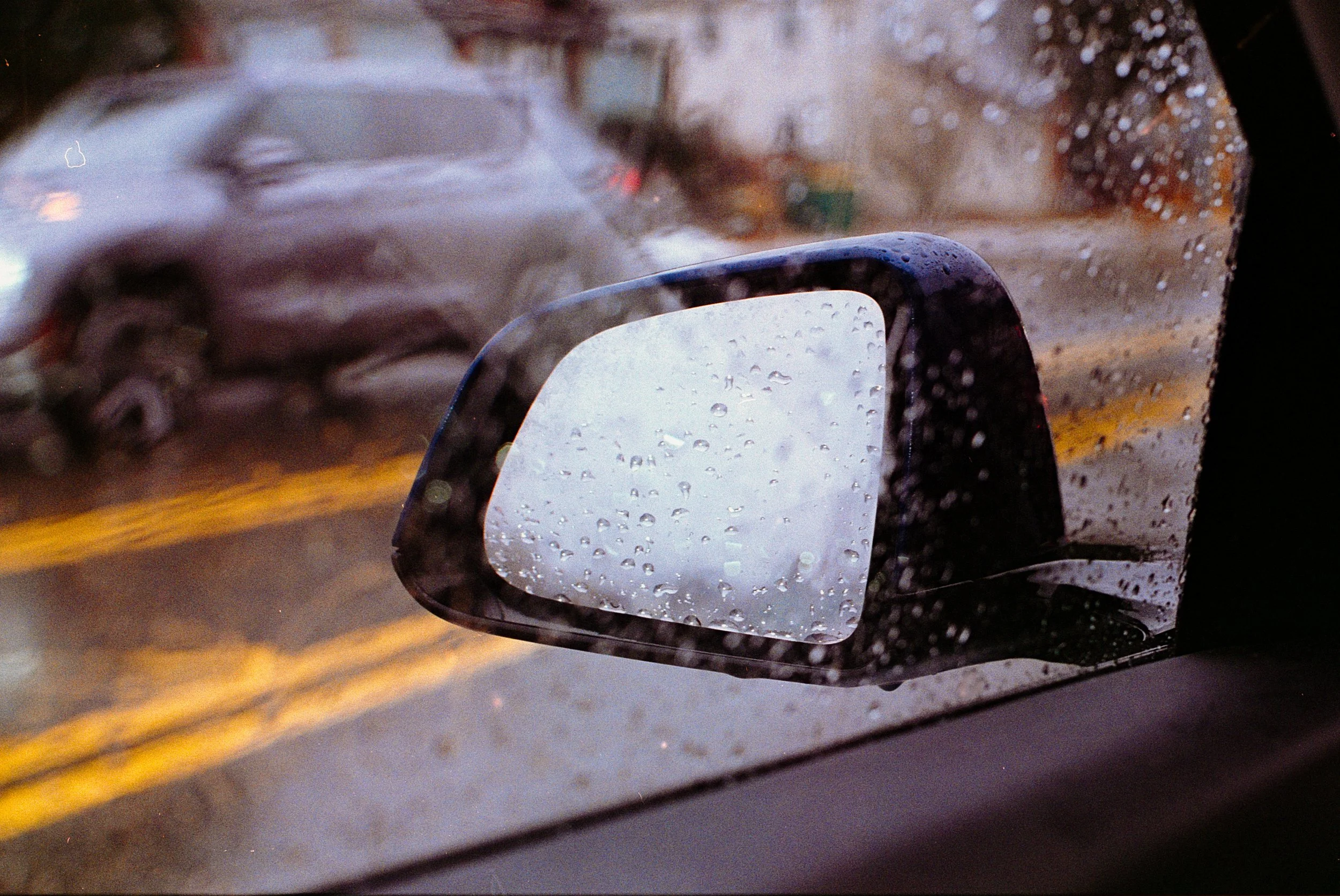 Close-up of a side car mirror of a car with raindrops on it, showing a blurred view of another vehicle and street scene through the rain-covered glass.