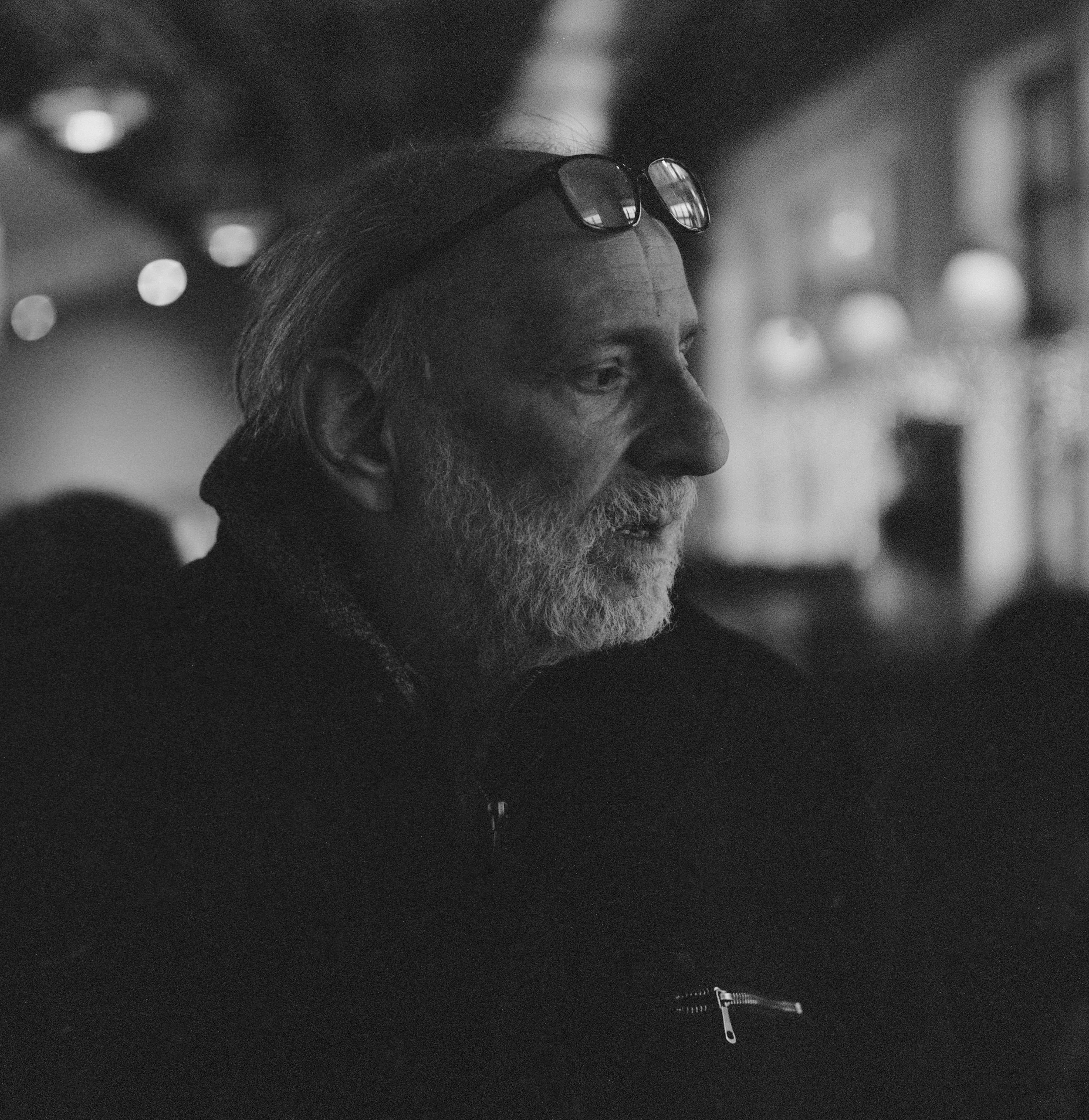 A black and white photo of an elderly man with a beard, glasses on his head, and short hair, sitting indoors in what appears to be a dimly lit setting, possibly a cafe or restaurant, with blurred lights and background details.