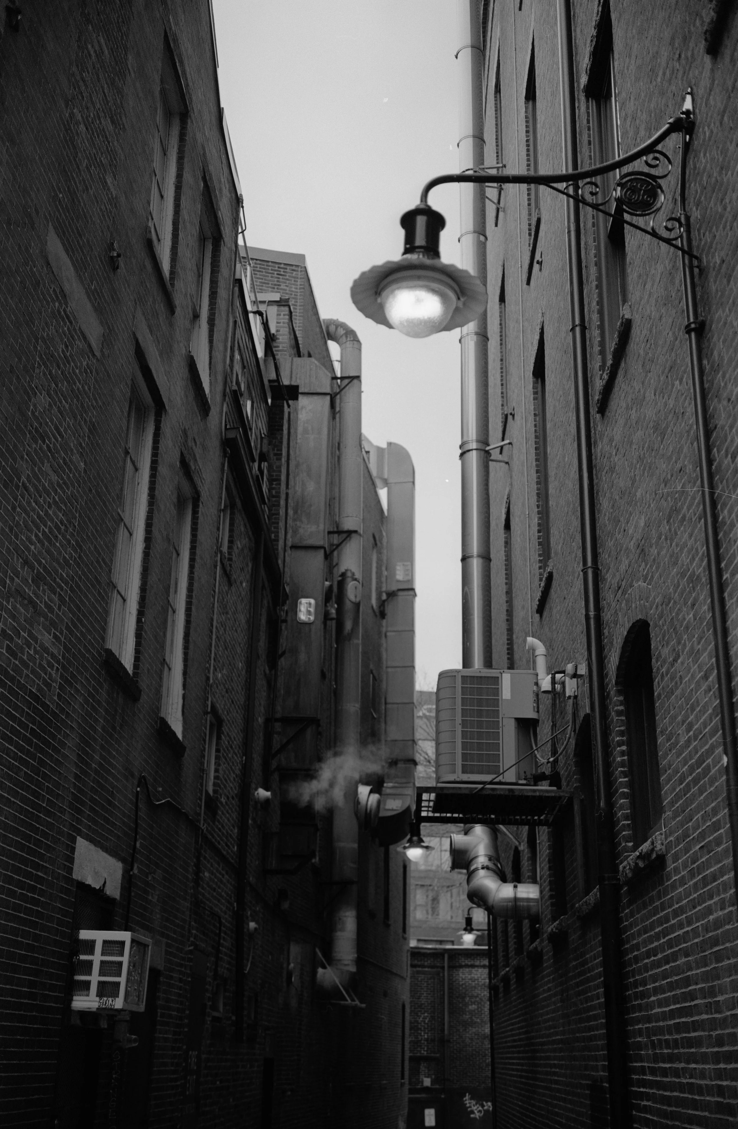 Black and white photo of a narrow city alleyway with brick buildings on each side, industrial ducts and pipes along the walls, and street lamps hanging above.