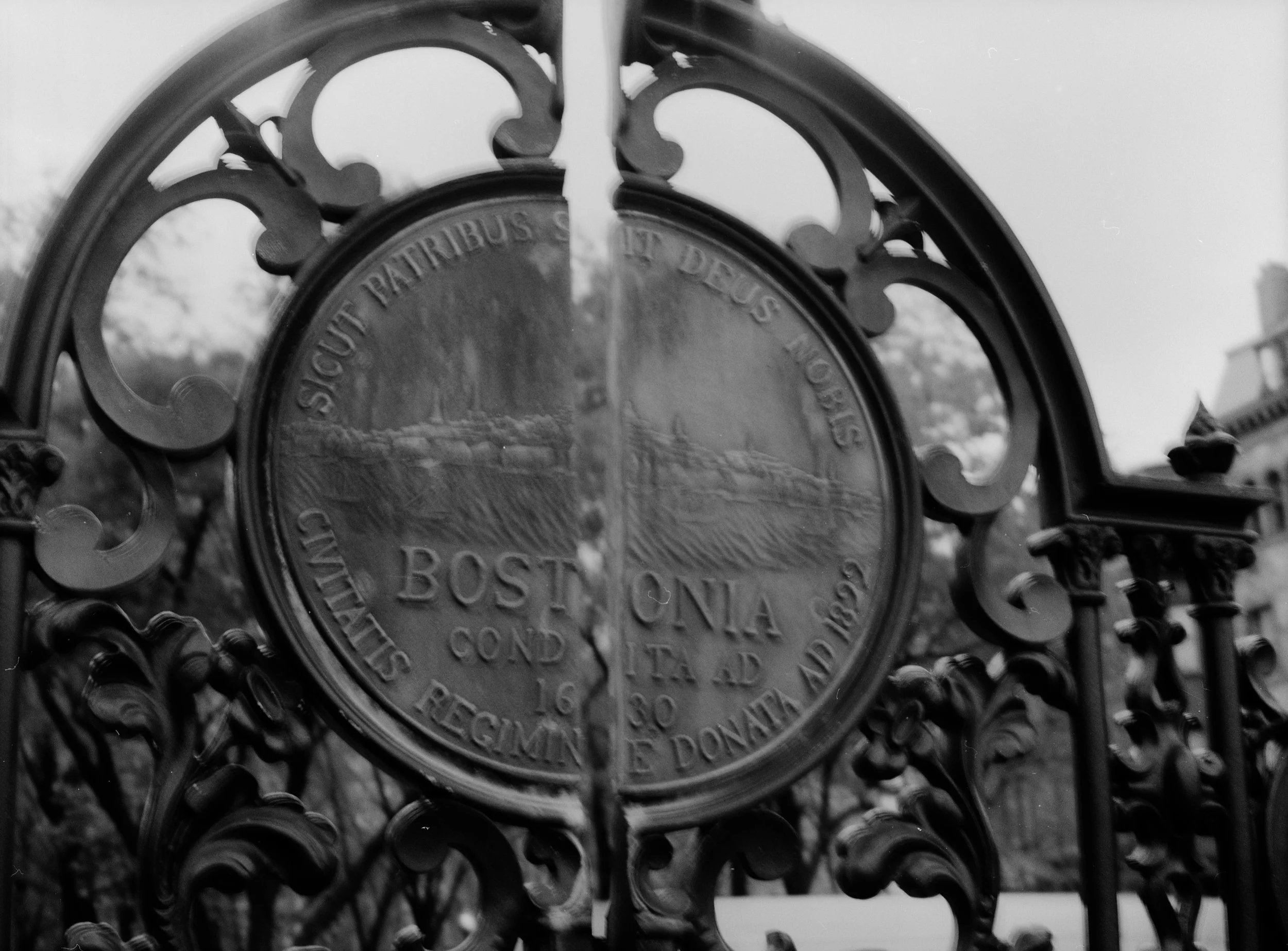 Close-up of an oval historical marker plaque on an ornate black metal fence, with a detailed cityscape and trees in the background.