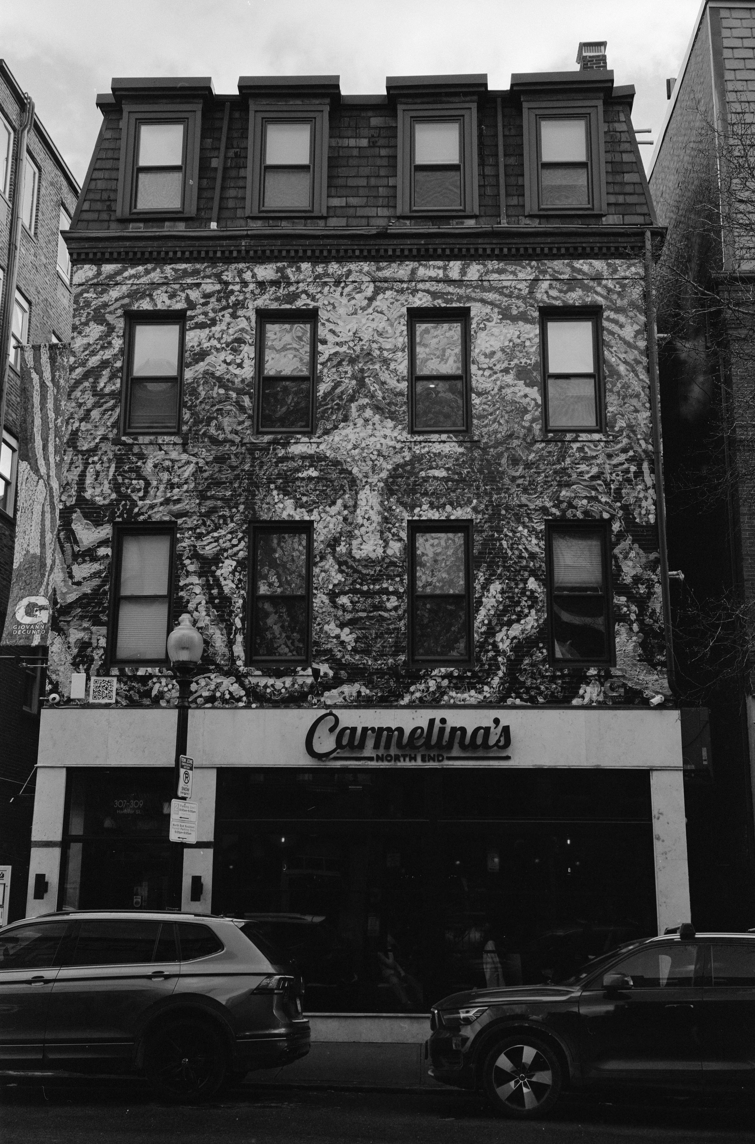 A multi-story building with a mural of a person's face painted on the upper floors. The building has a storefront on the ground level labeled 'Carmelina's North End.' Cars are parked in front of the building.