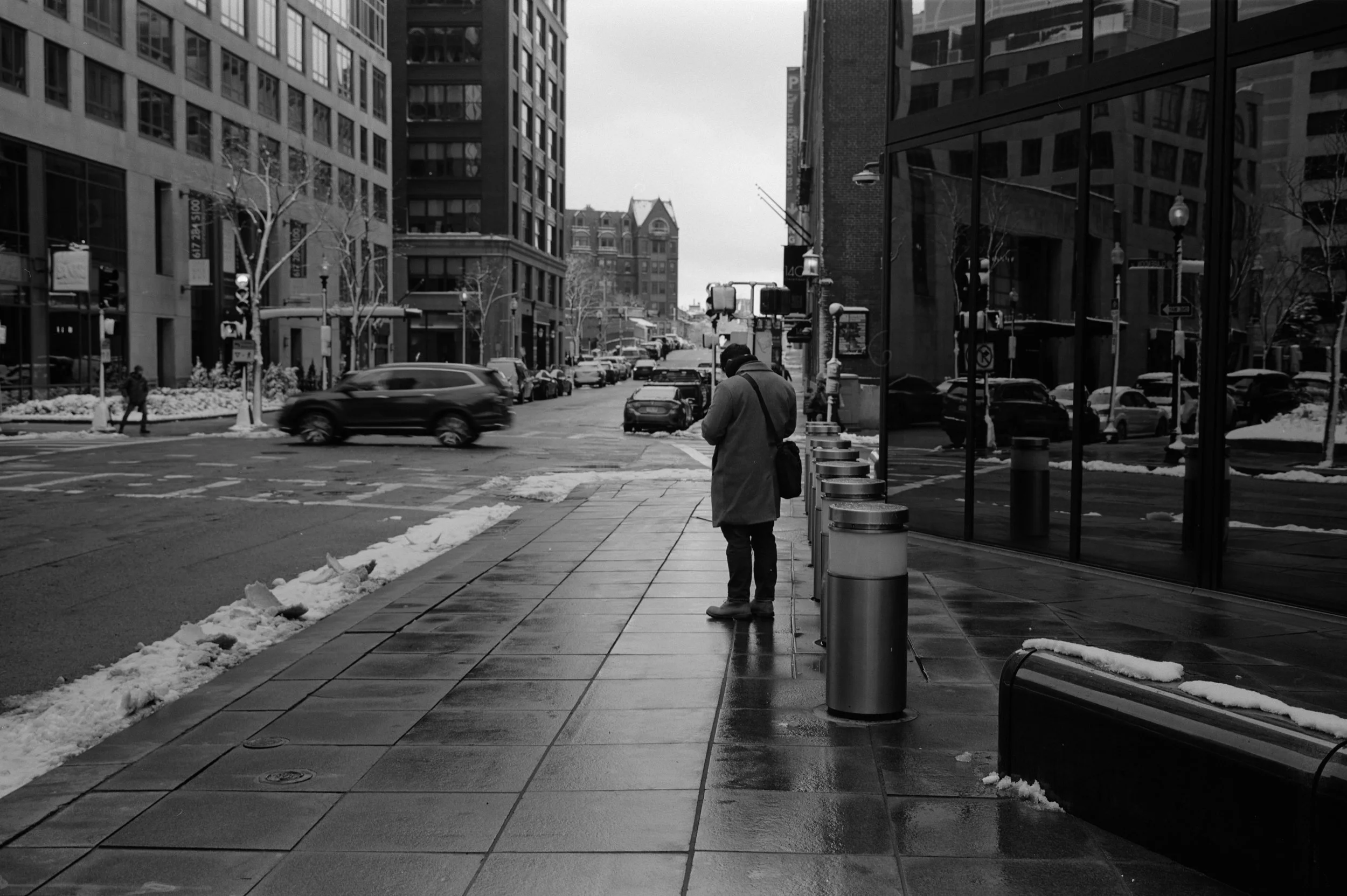 A person standing alone on the sidewalk of a city street on a snowy winter day, with buildings and cars in the background, and snow piled along the curb.