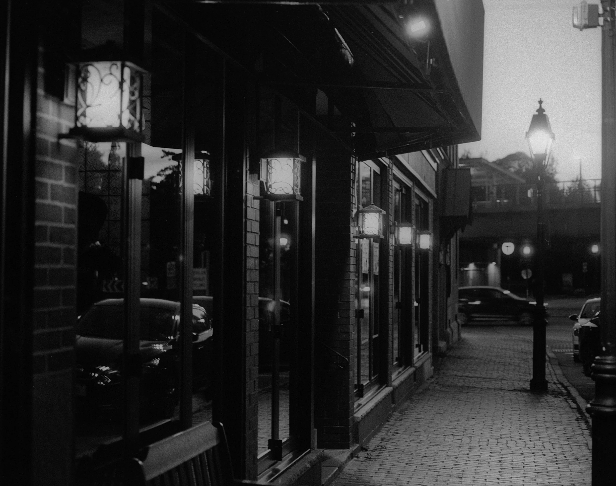 Nighttime street scene with illuminated lantern-style street lamps along a sidewalk next to a building with large windows and brick walls. Cars are parked and driving by, and a traffic light is visible in the background.