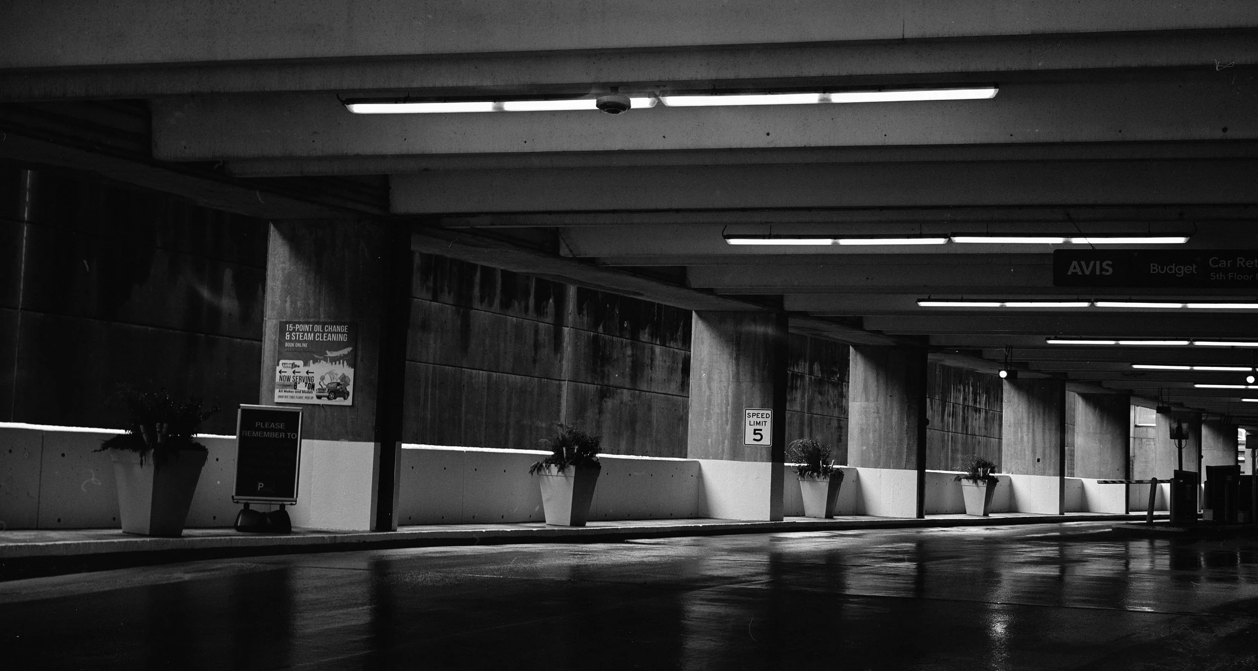 An empty parking garage with concrete walls and ceiling, illuminated by fluorescent ceiling lights, white planters with plants lined up along the wall, a sign indicating a speed limit of 5, and a small blackboard sign in the foreground.
