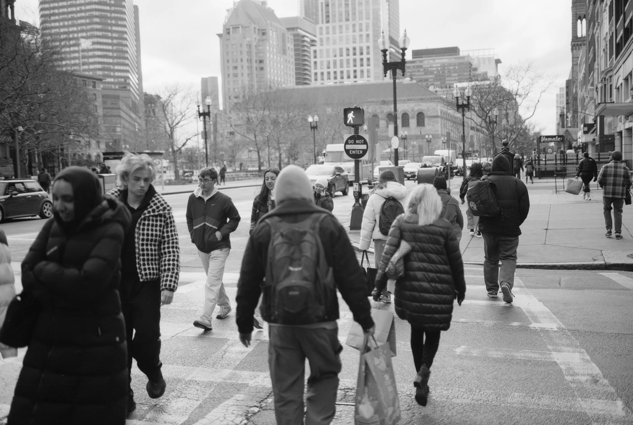 People crossing a city street at a crosswalk with tall buildings and trees in the background, black and white photo.