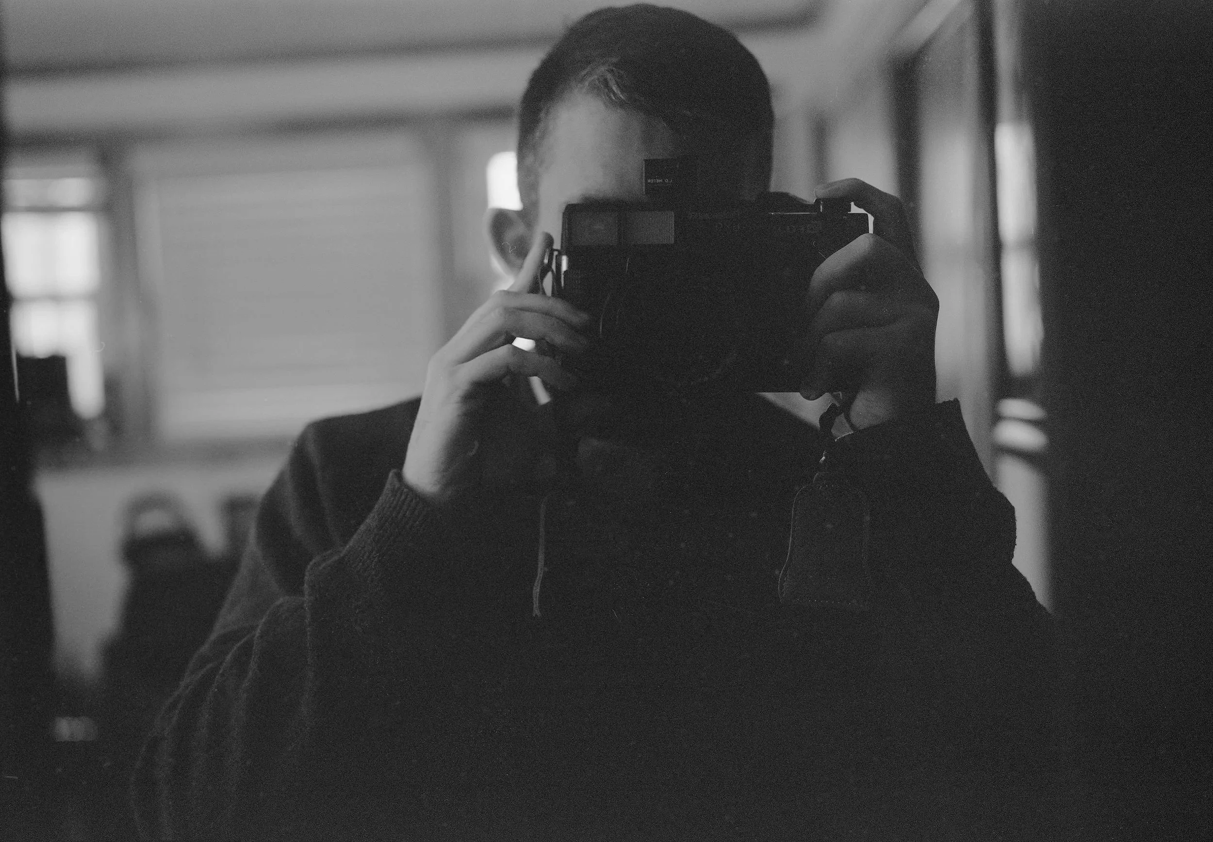 A person taking a black and white photo of themselves in a mirror, holding a camera up to their face, with a background of windows and other people in an indoor space.