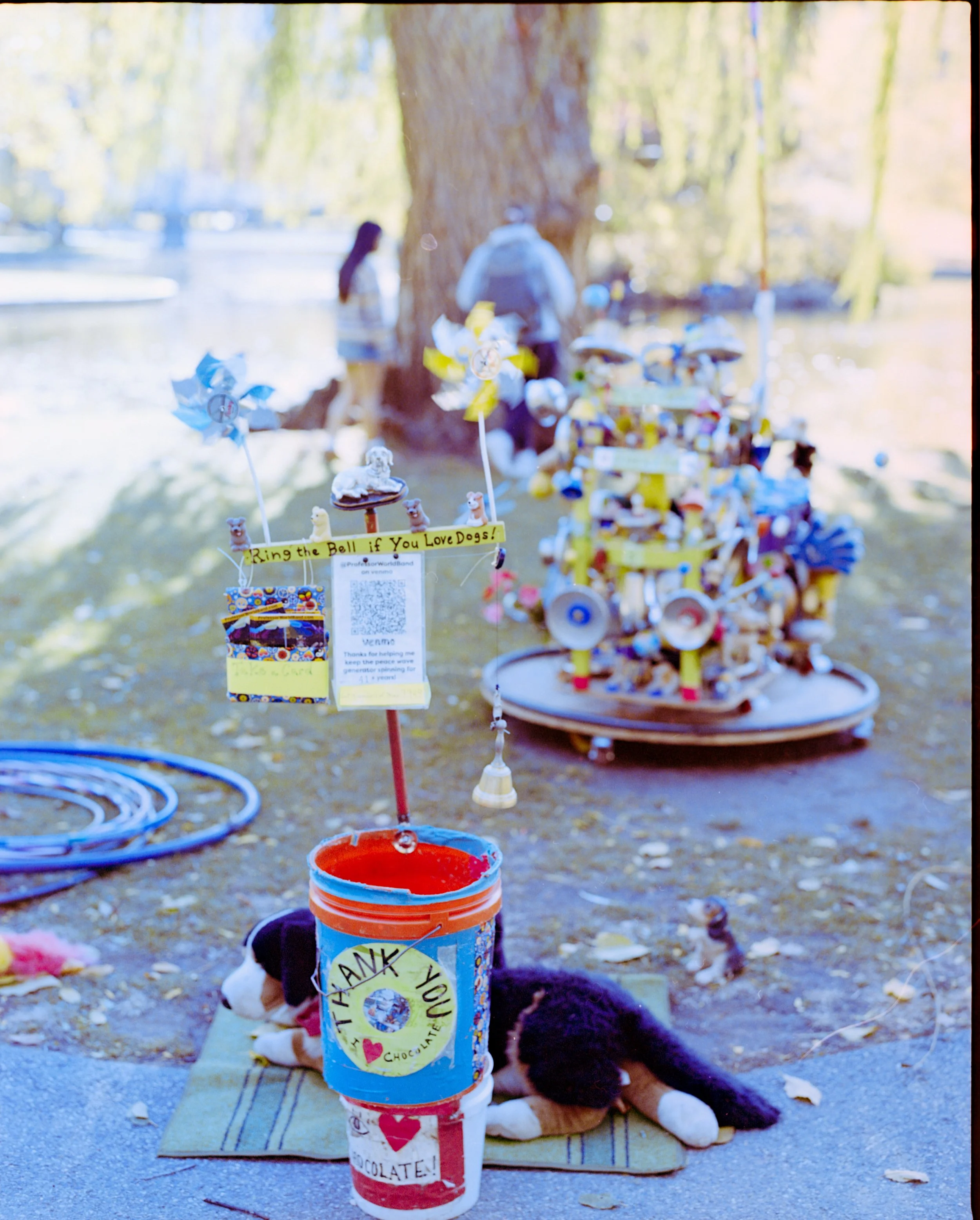 Outdoor scene with a donation bucket decorated with a thank you sticker, surrounded by small toys and stuffed animals, and a sign encouraging donations for dogs, with people and a large tree in the background, and a small dog plush on the ground in f