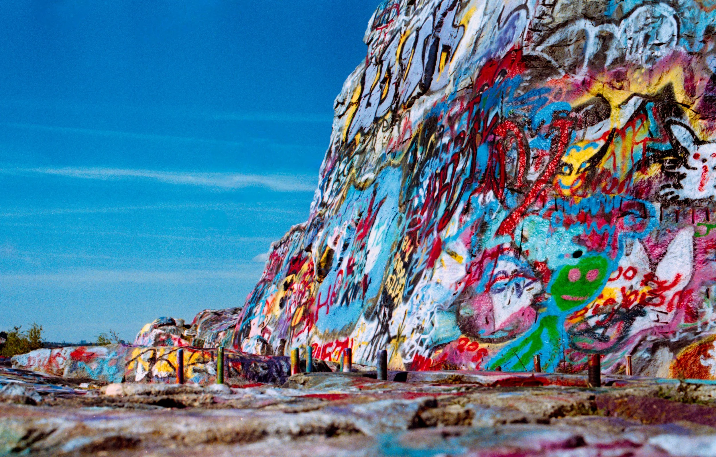 A colorful graffiti mural painted on a large rock face, with various abstract shapes, drawings, and writings, against a clear blue sky.